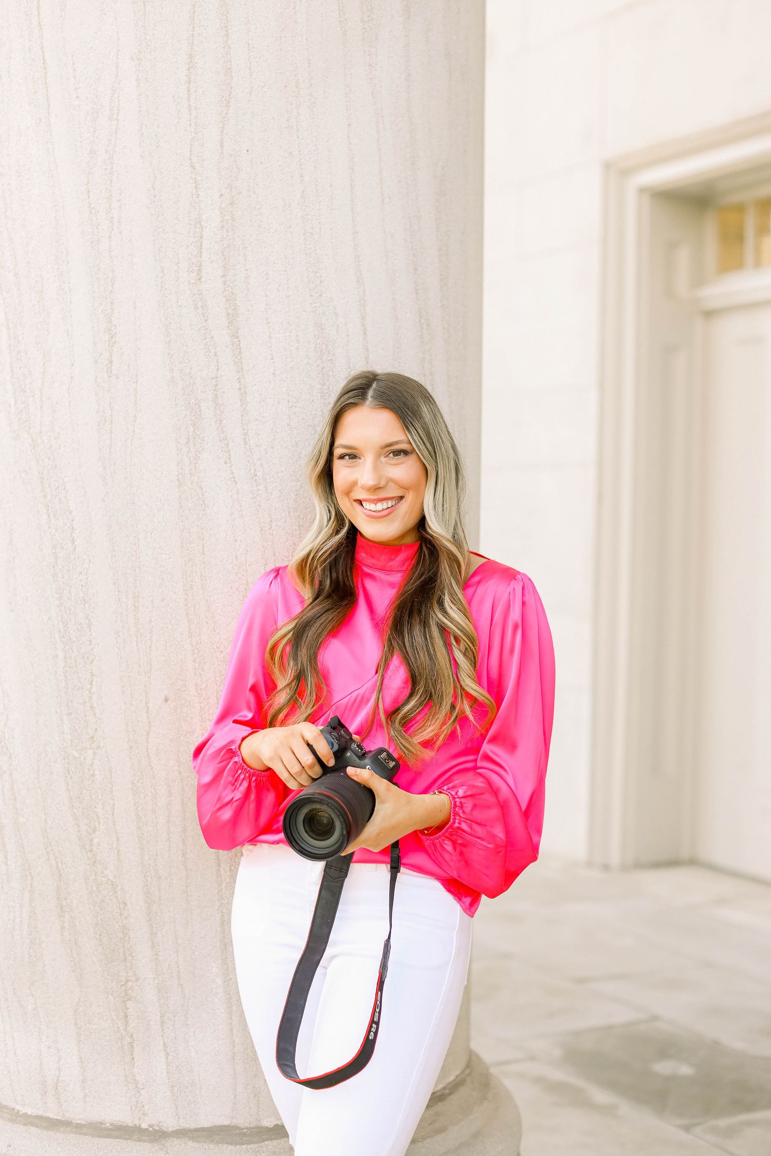 Professional photographer in vibrant pink jacket demonstrates camera gear in white studio space.