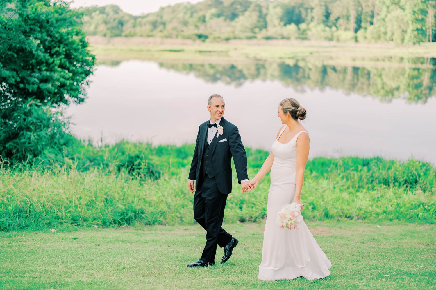 Bride and groom hold hands near a serene lake, surrounded by lush greenery, on their wedding day.