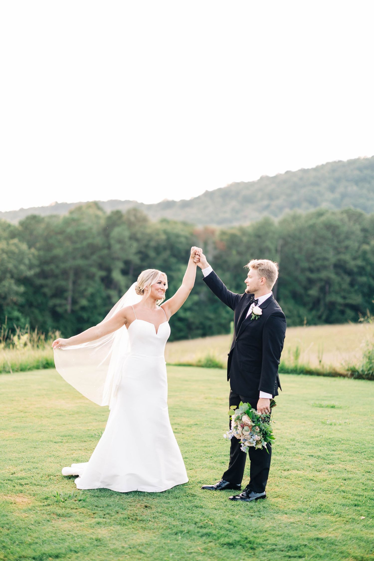 Newlyweds dance joyfully on green lawn with mountain vista behind them.