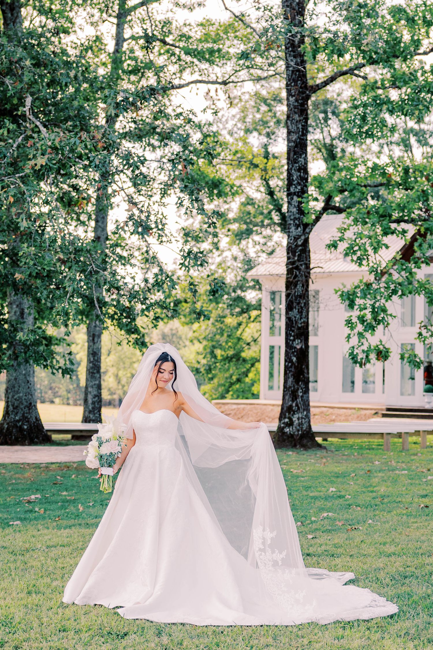 Bride in flowing white dress and veil standing on lawn with trees in background.