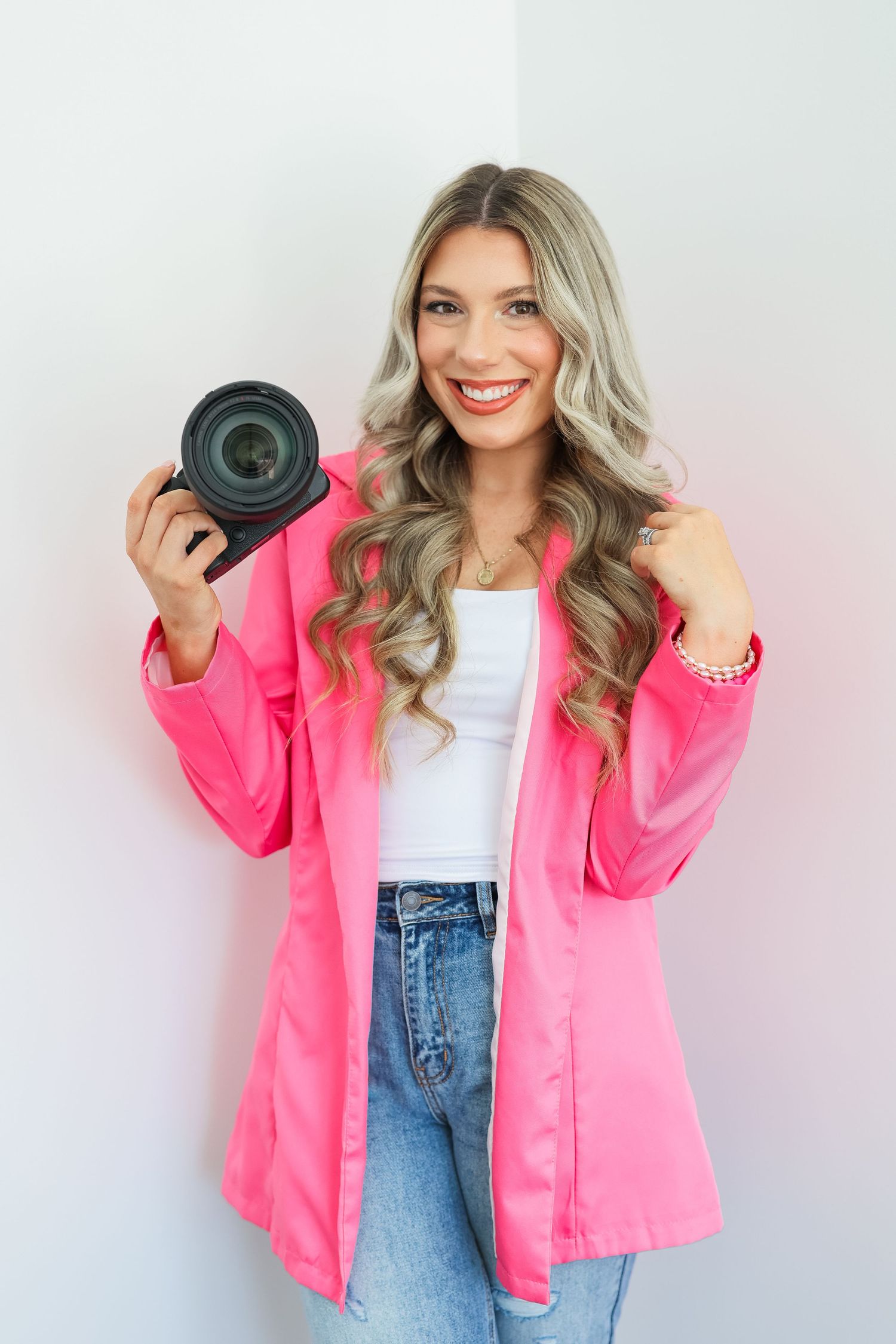 Creative portrait of photographer holding camera lens while wearing bright pink jacket and denim.