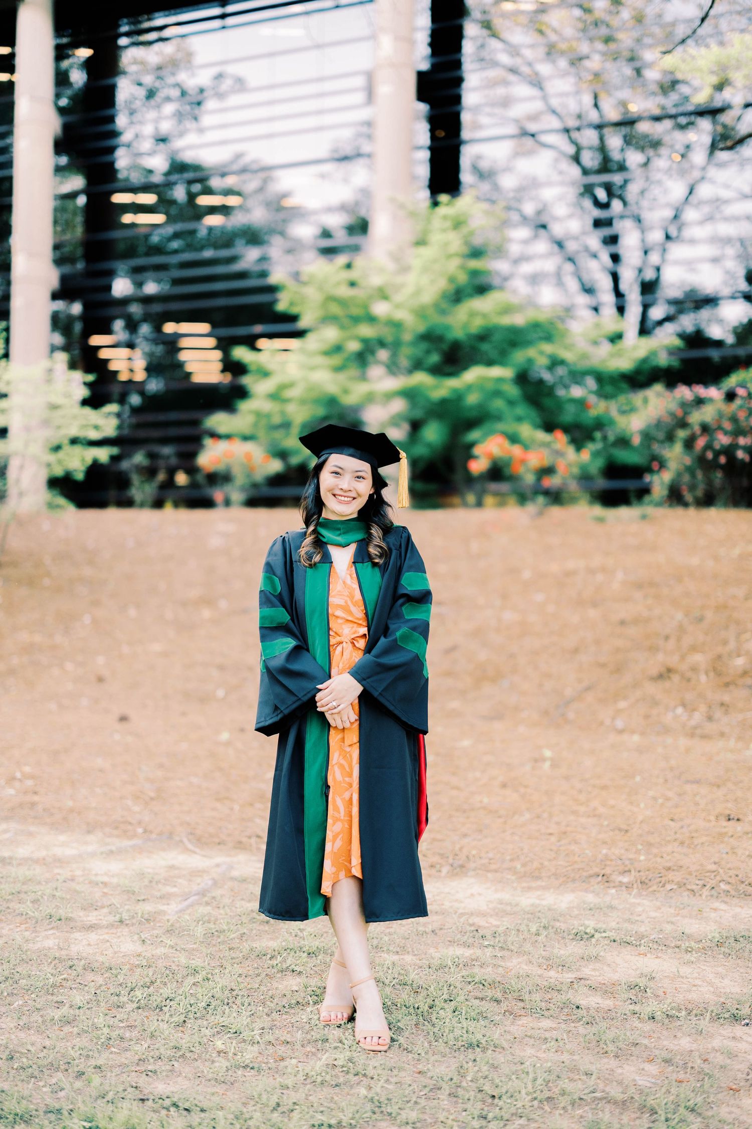A graduate in green academic regalia poses on grass for graduation photos.