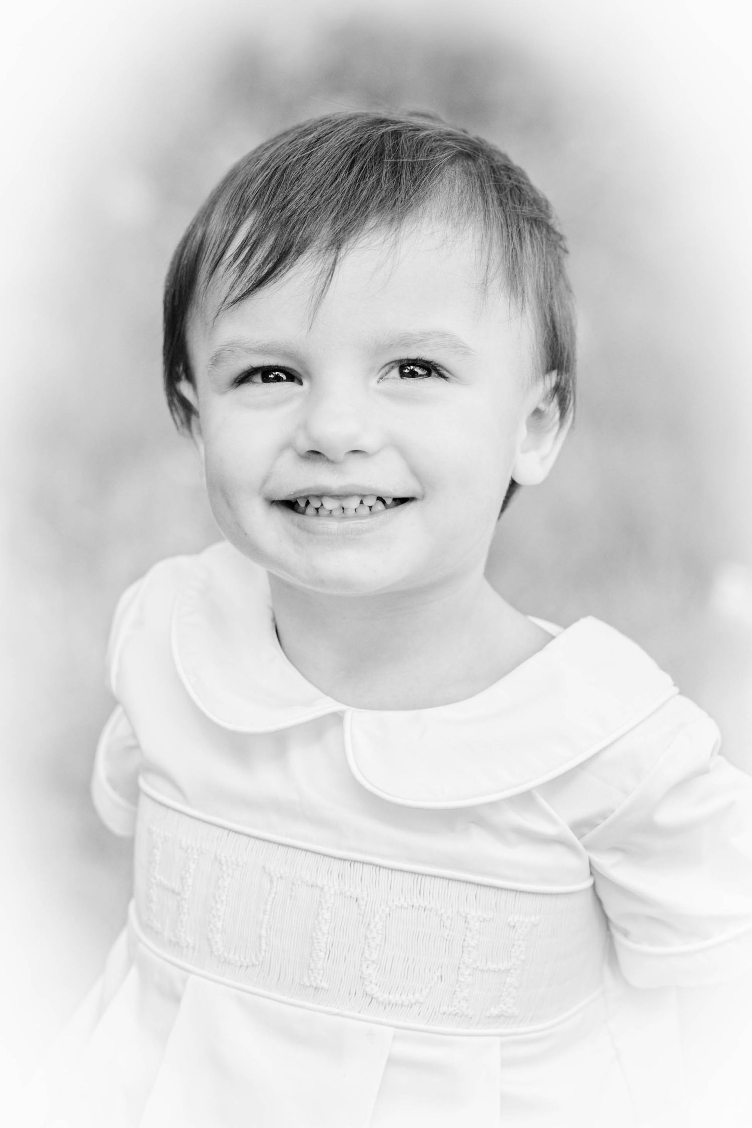Black and white portrait of a smiling toddler in a white smocked dress, soft bokeh background.