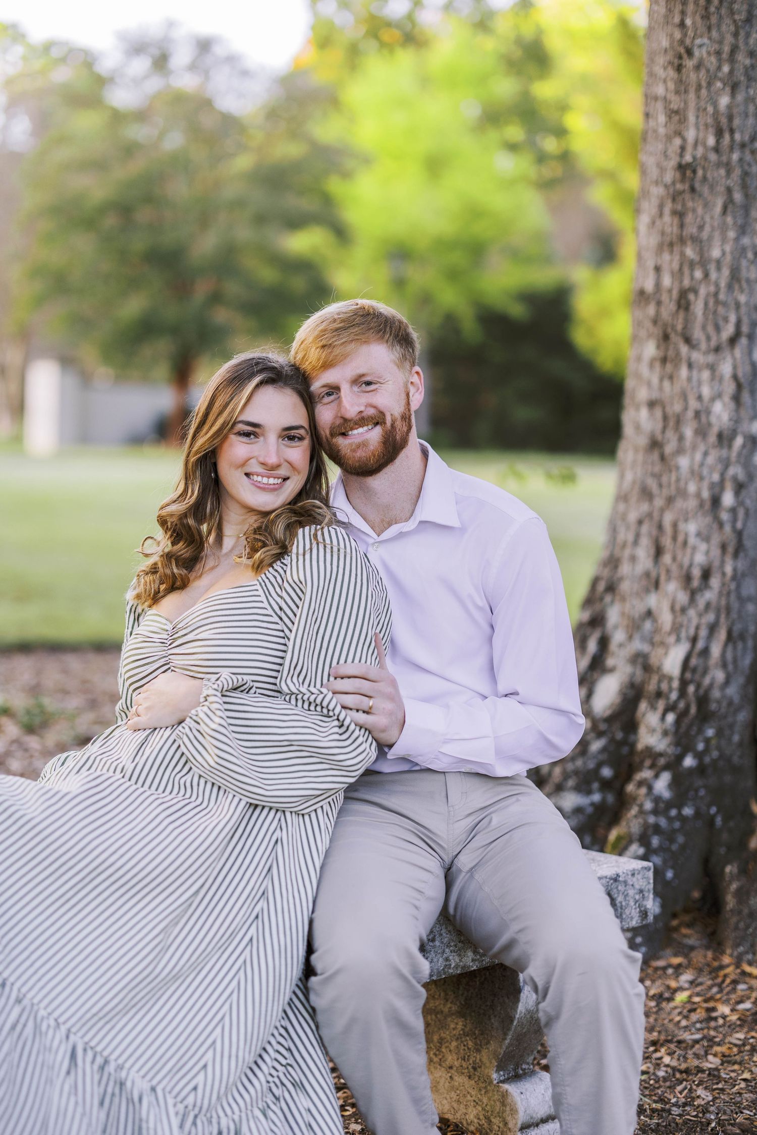 A couple sits against a tree trunk sharing a tender moment in a park setting.