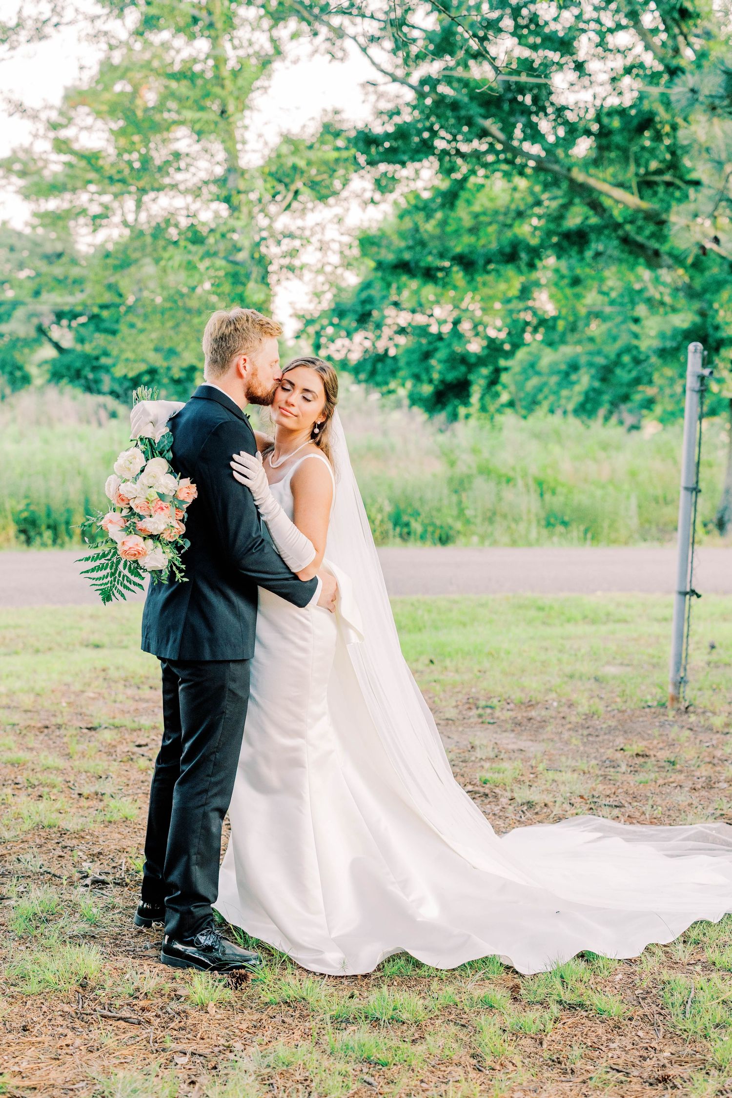 A romantic outdoor wedding portrait with flowing veil and pink bouquet against green foliage.