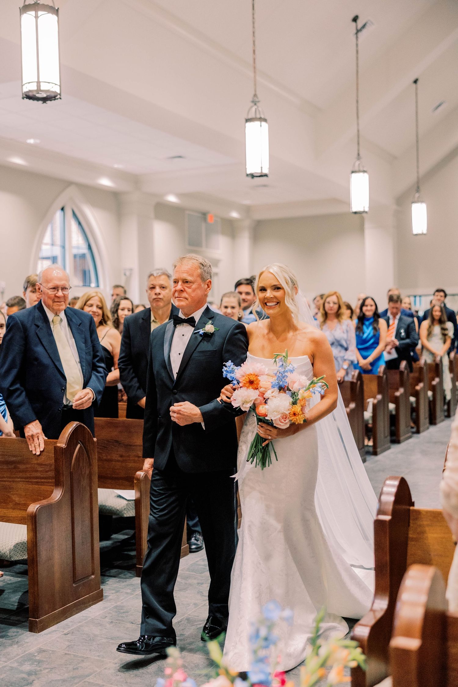 Wedding ceremony processional in a bright church with guests watching from wooden pews.
