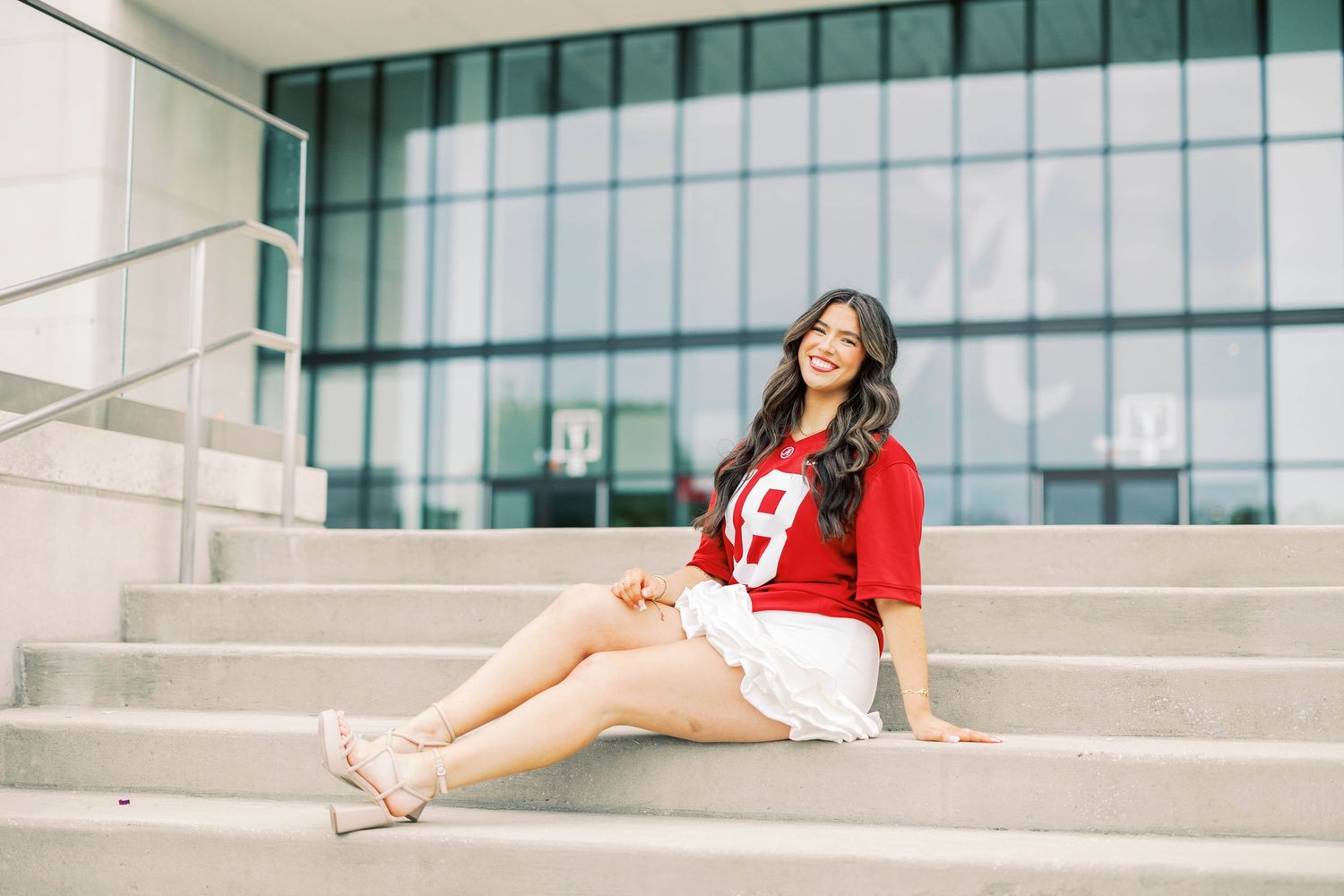 Person in graduation attire posing on white concrete steps in front of glass building.