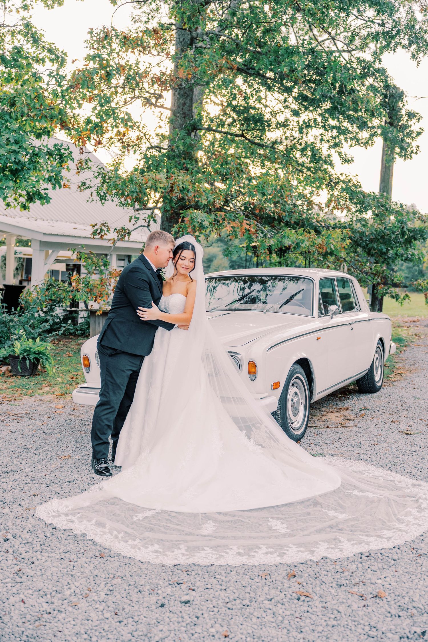 Wedding couple posing by vintage white car under large trees.