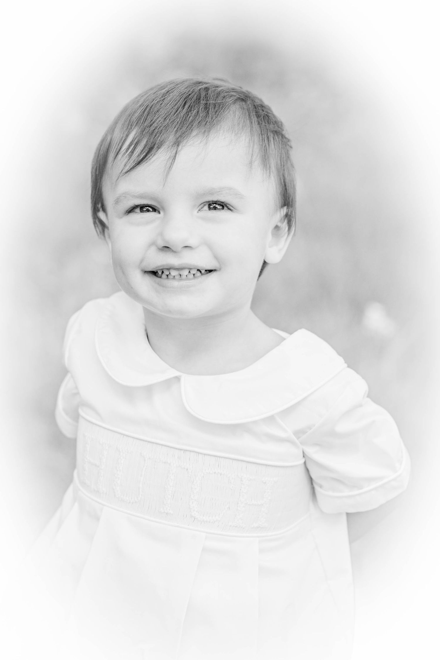 Black and white portrait of a smiling toddler in a white smocked dress with peter pan collar.