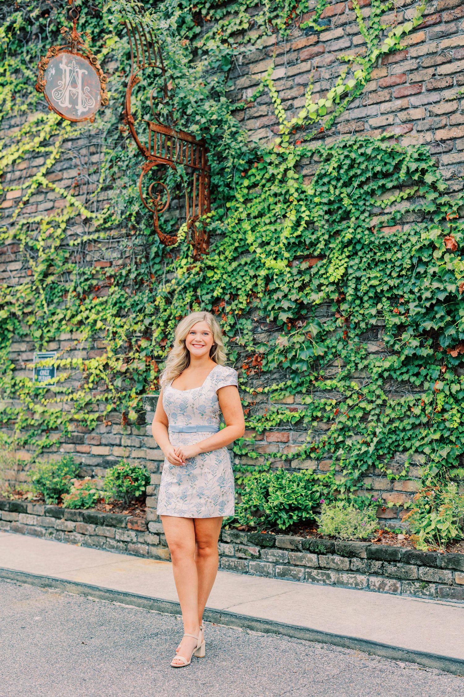 Smiling blonde woman in floral mini dress standing on sidewalk before ivy-covered brick wall with decorative mirror.