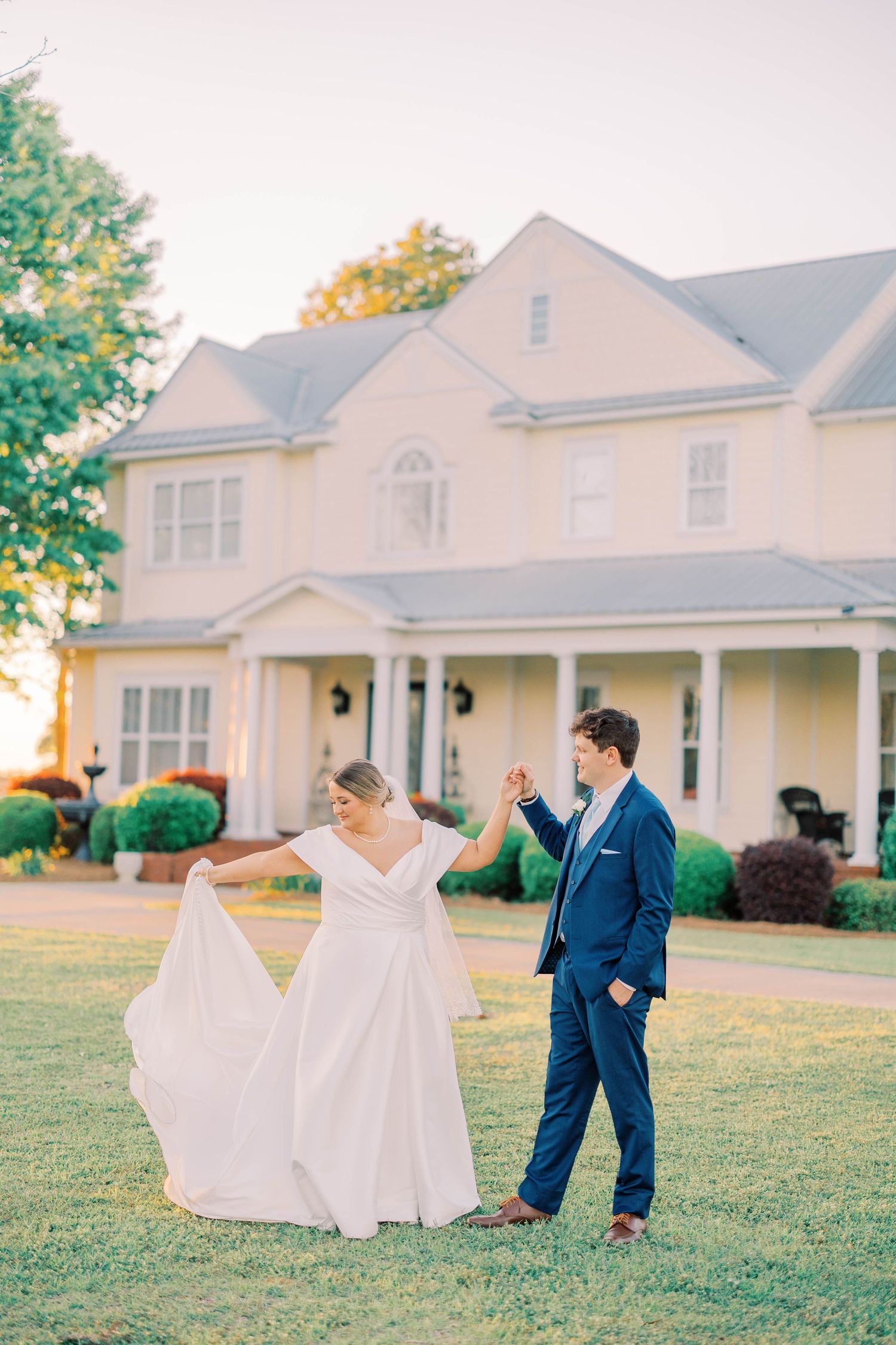 Newlyweds dance playfully on a grassy lawn with a white house in the background.
