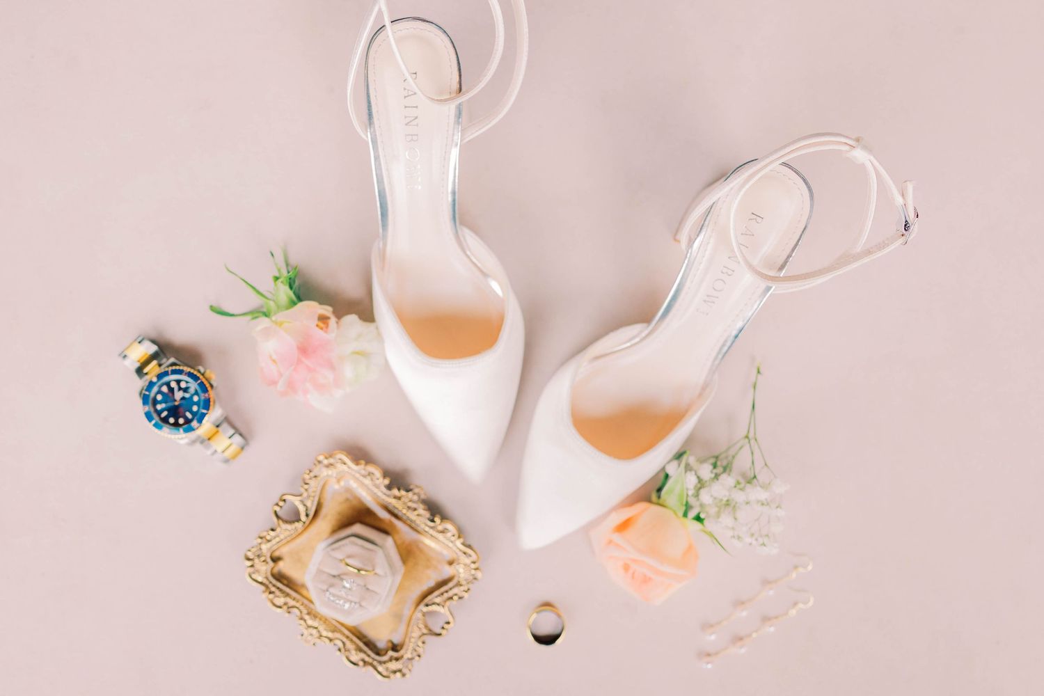 White bridal heels with ankle straps styled with floral accents, a blue watch, and a gold ring on a pink background.