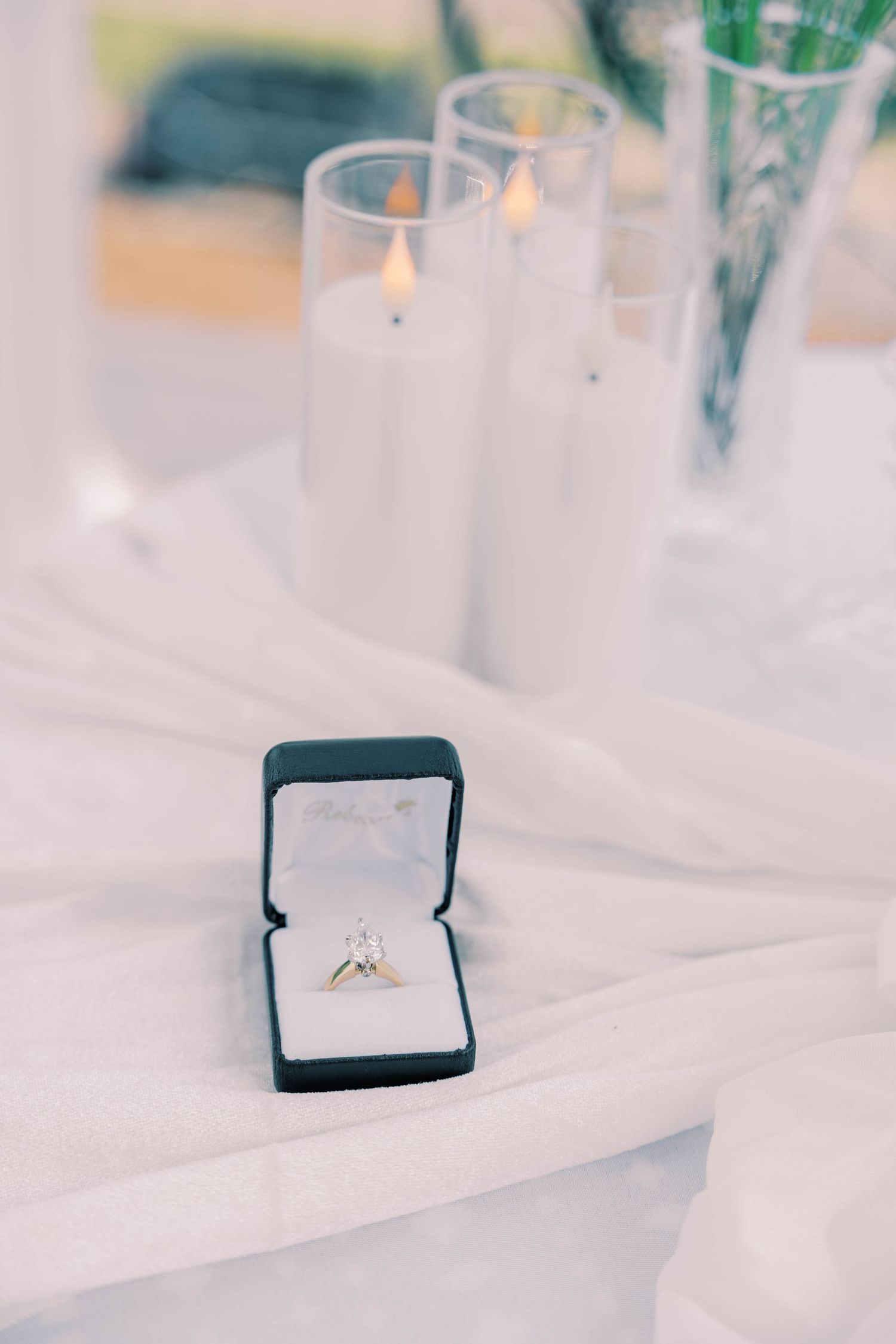 A delicate engagement ring sits in a navy blue velvet box with soft lighting and candles in the background.