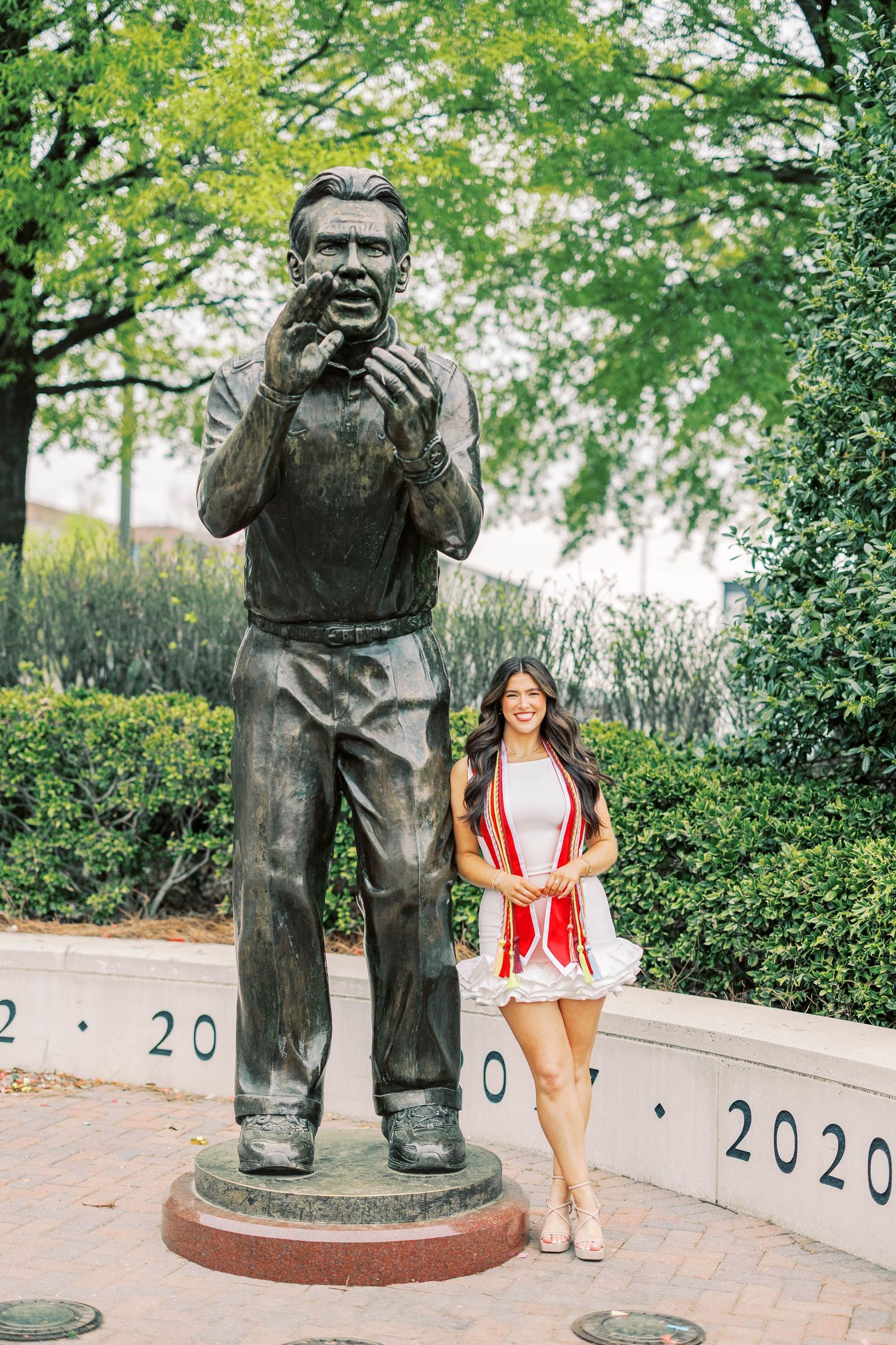 Bronze statue and person in red outfit posing together on brick pathway with greenery background.