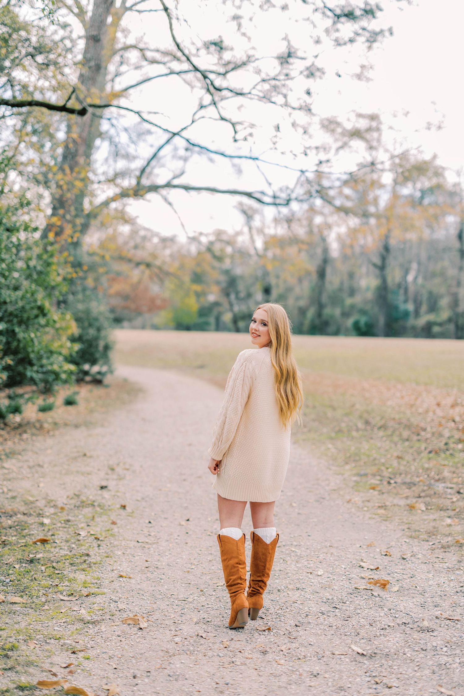 Woman in beige sweater dress and brown boots walking on autumn path through trees.
