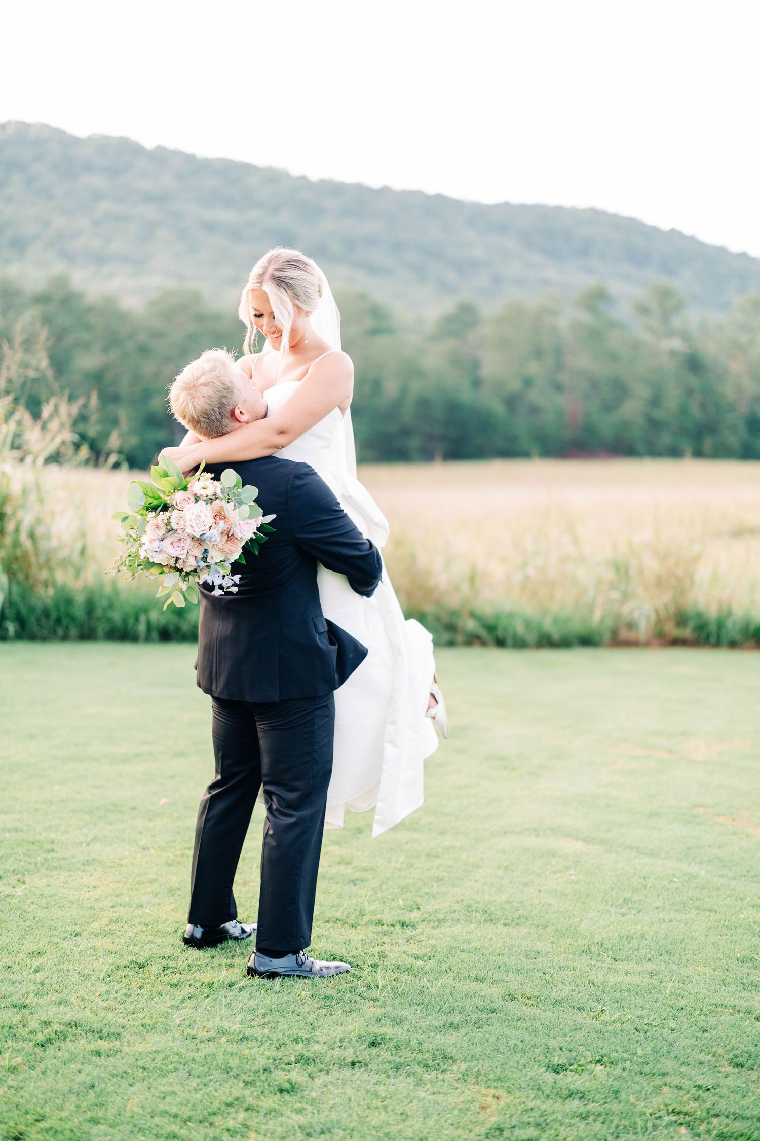 Romantic wedding couple embraces on grassy hill with mountains in background.