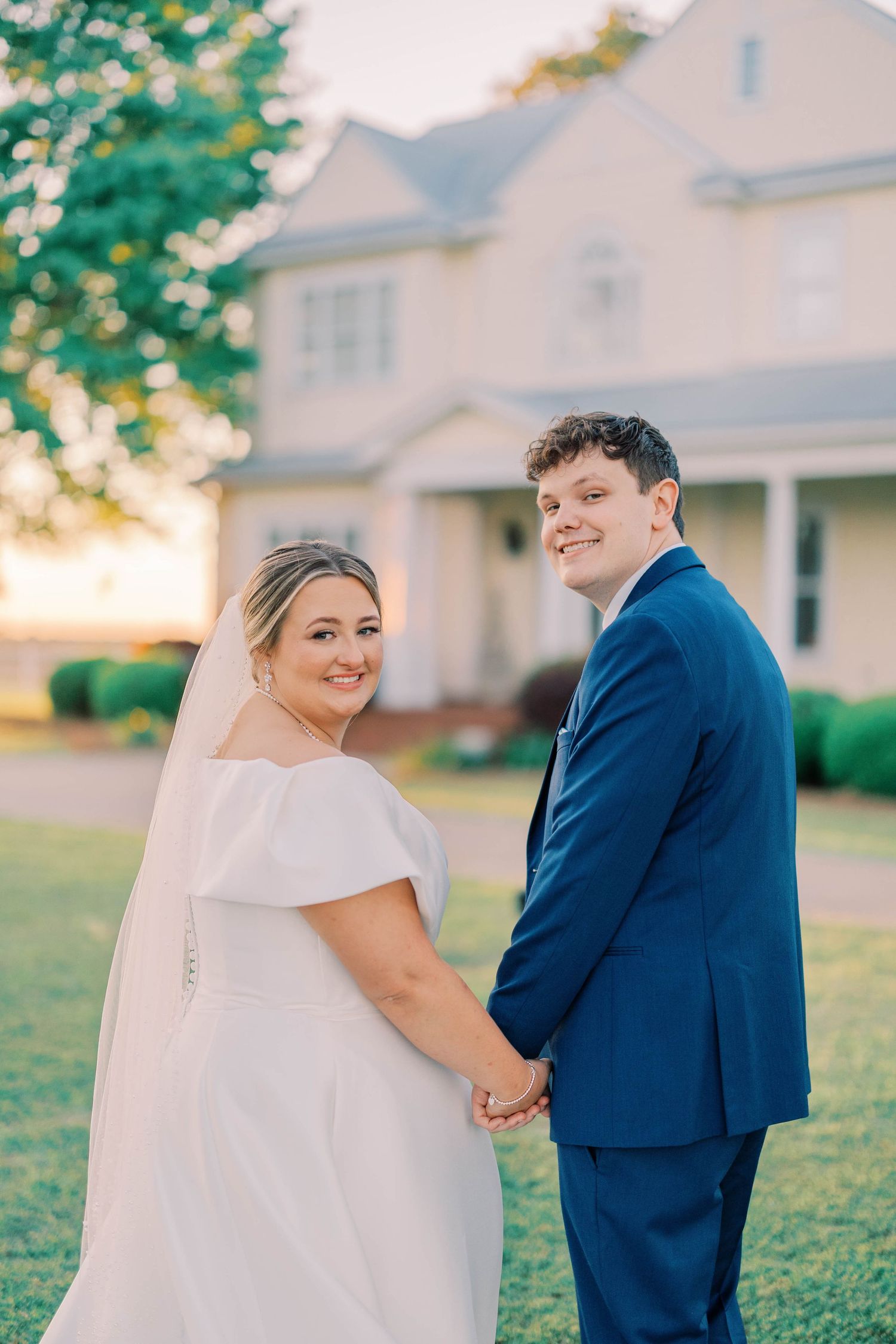 A couple in wedding attire shares a romantic moment on a lawn in front of a white house during golden hour.