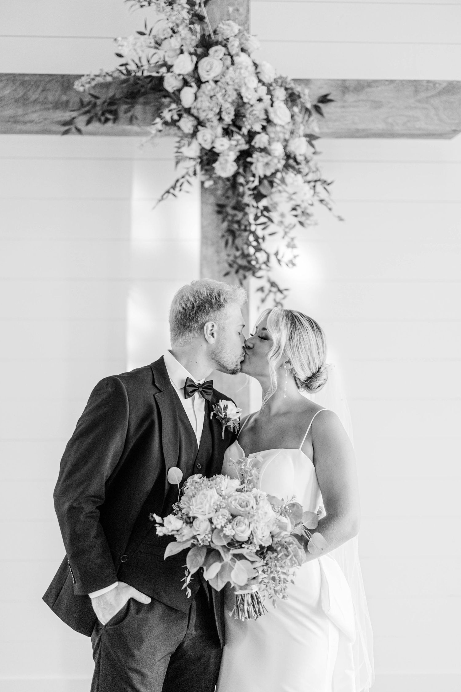 Black and white photo of couple sharing intimate moment under floral arrangement.