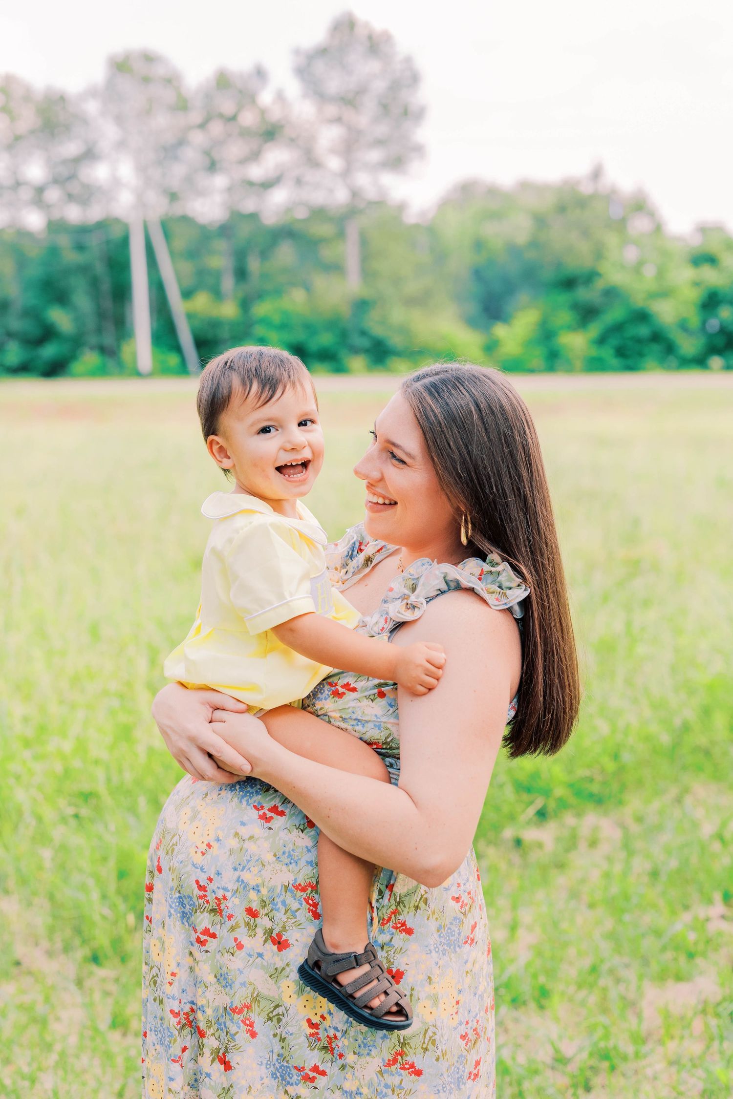 A mother in a floral dress holds her laughing toddler in a yellow shirt outdoors in a green field.