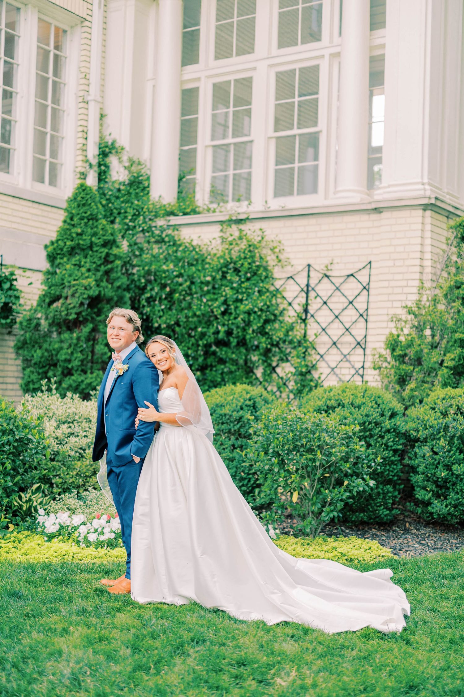 Bride in white gown and groom in blue suit pose back-to-back in lush garden outside elegant venue.