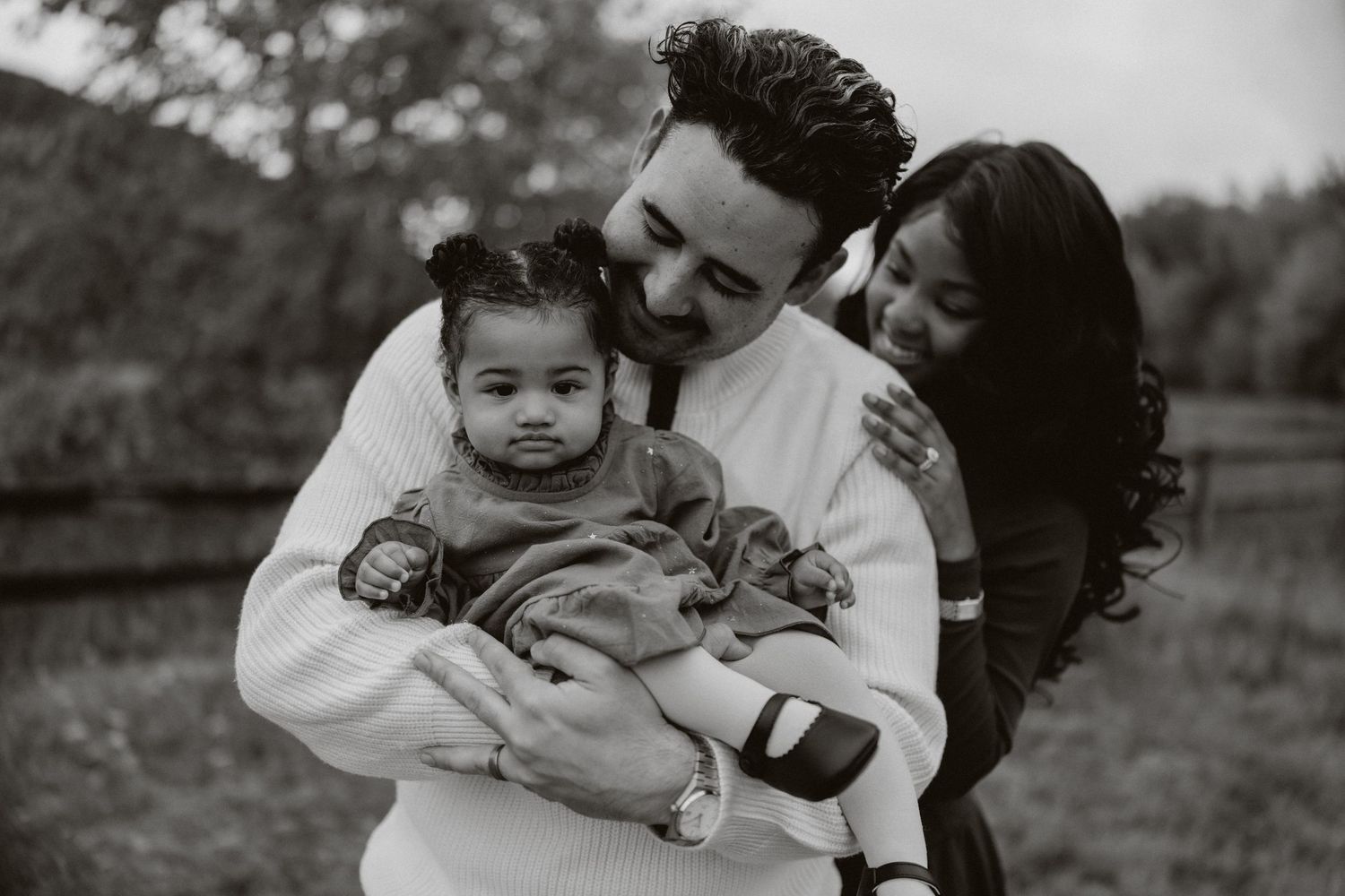 A black and white photograph of a family embracing outdoors in a natural setting.