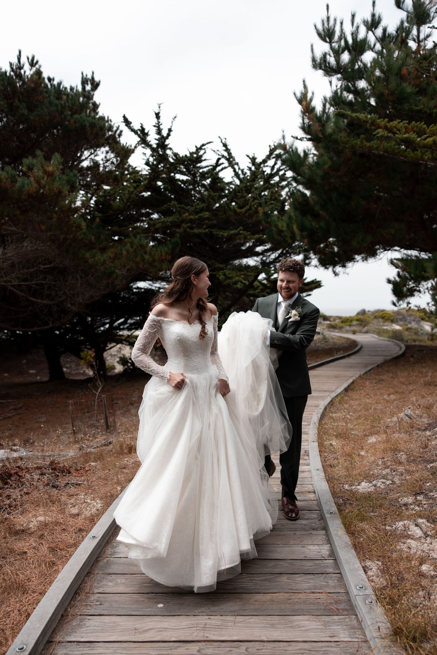 A couple walks together on a wooden boardwalk path through evergreen trees on their wedding day.