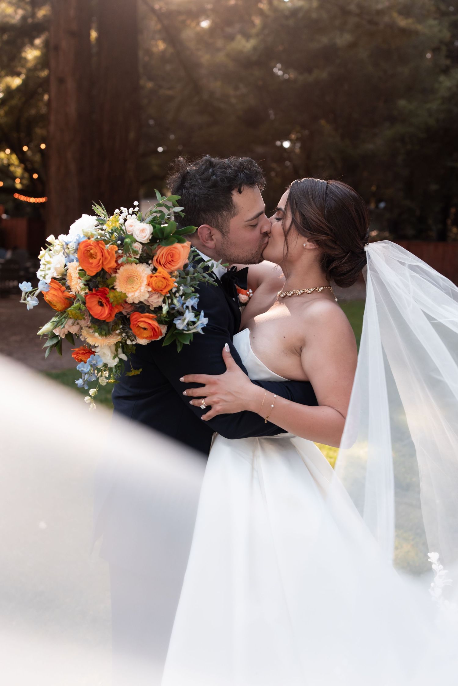A romantic wedding moment captured in a forest setting with orange floral bouquet and flowing white veil.