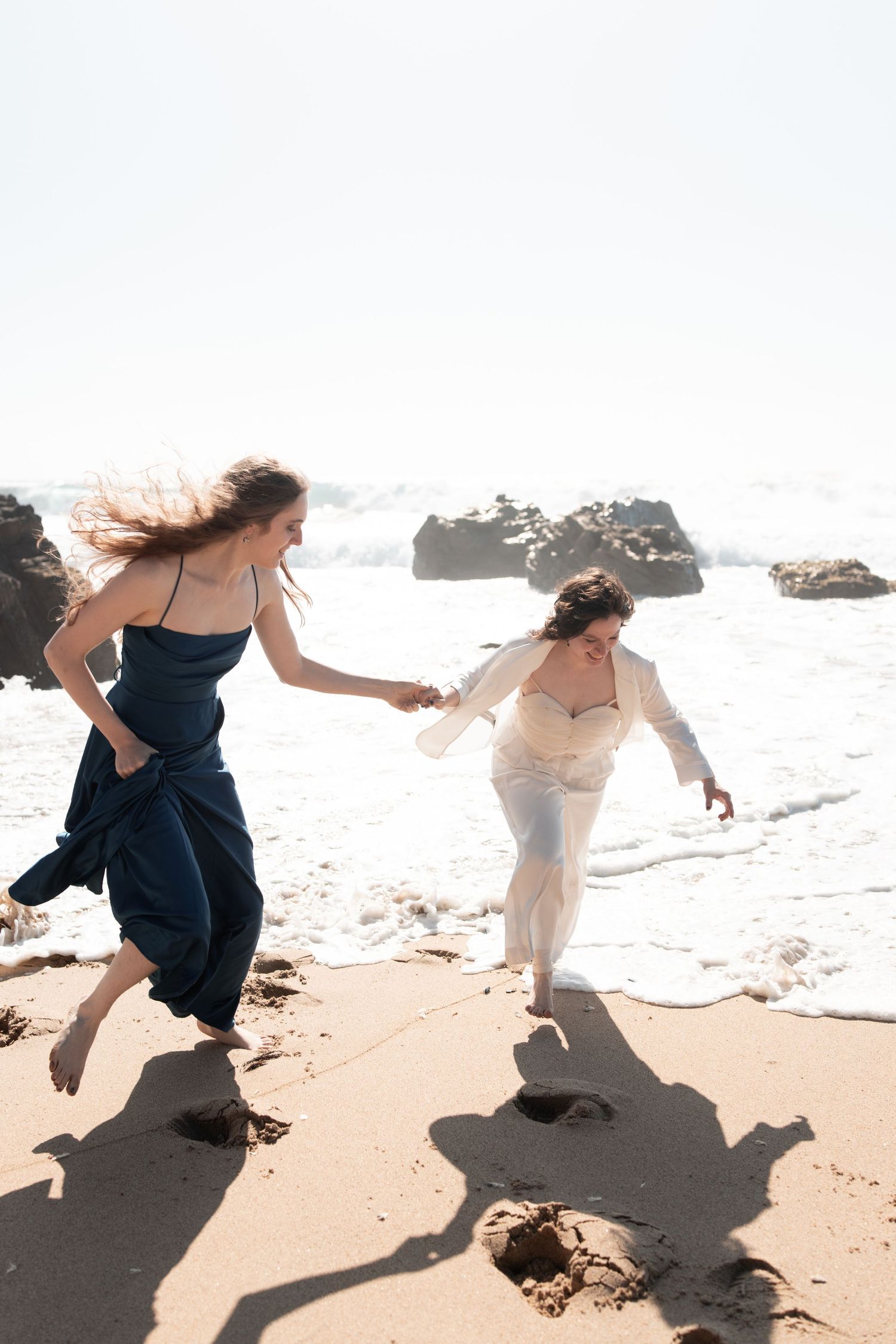 Two people in flowing dresses dance and leap gracefully along a sunlit beach shoreline.