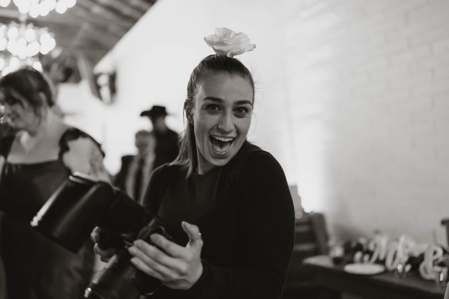 Black and white photo of a laughing woman balancing a flower on her head while holding a camera at an event.