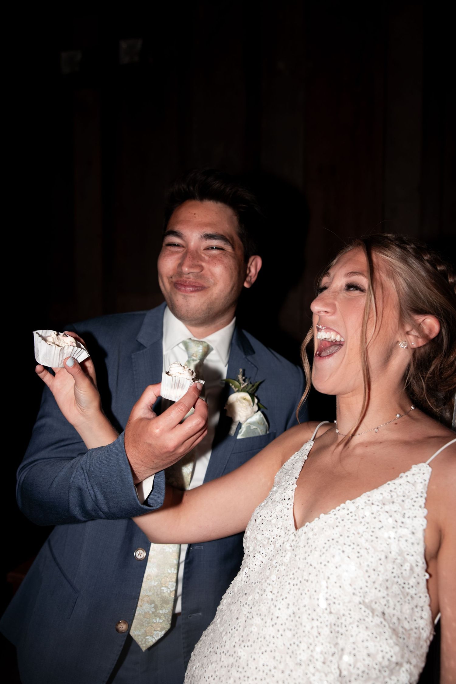 Bride and groom laughing joyfully during wedding cake cutting ceremony, groom in blue suit, bride in white gown.