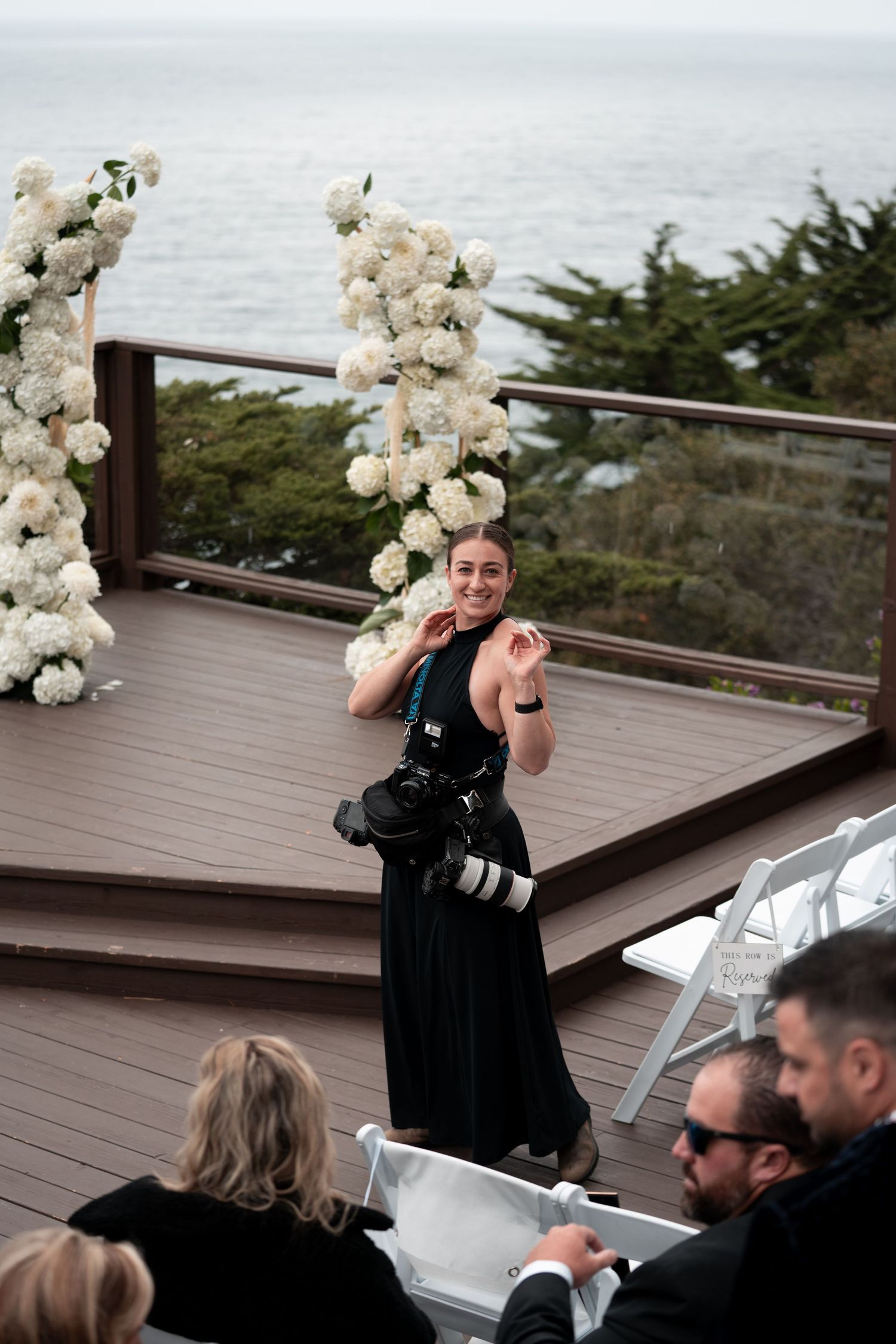 A smiling female photographer in a black dress carries a camera at an outdoor wedding ceremony with white floral arch by the ocean.
