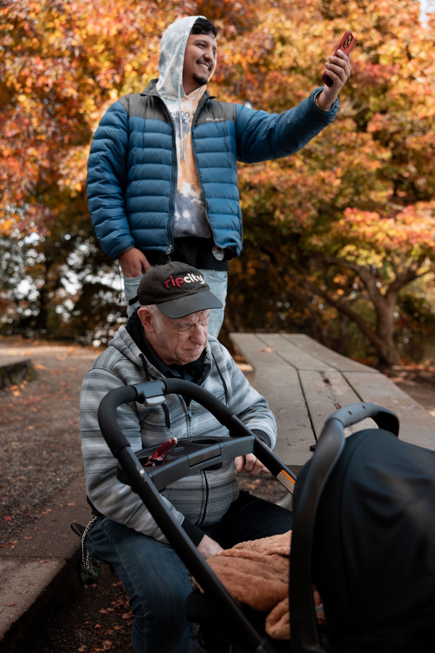 Person in blue puffer jacket and grey hoodie relaxing on wooden bench surrounded by autumn foliage.