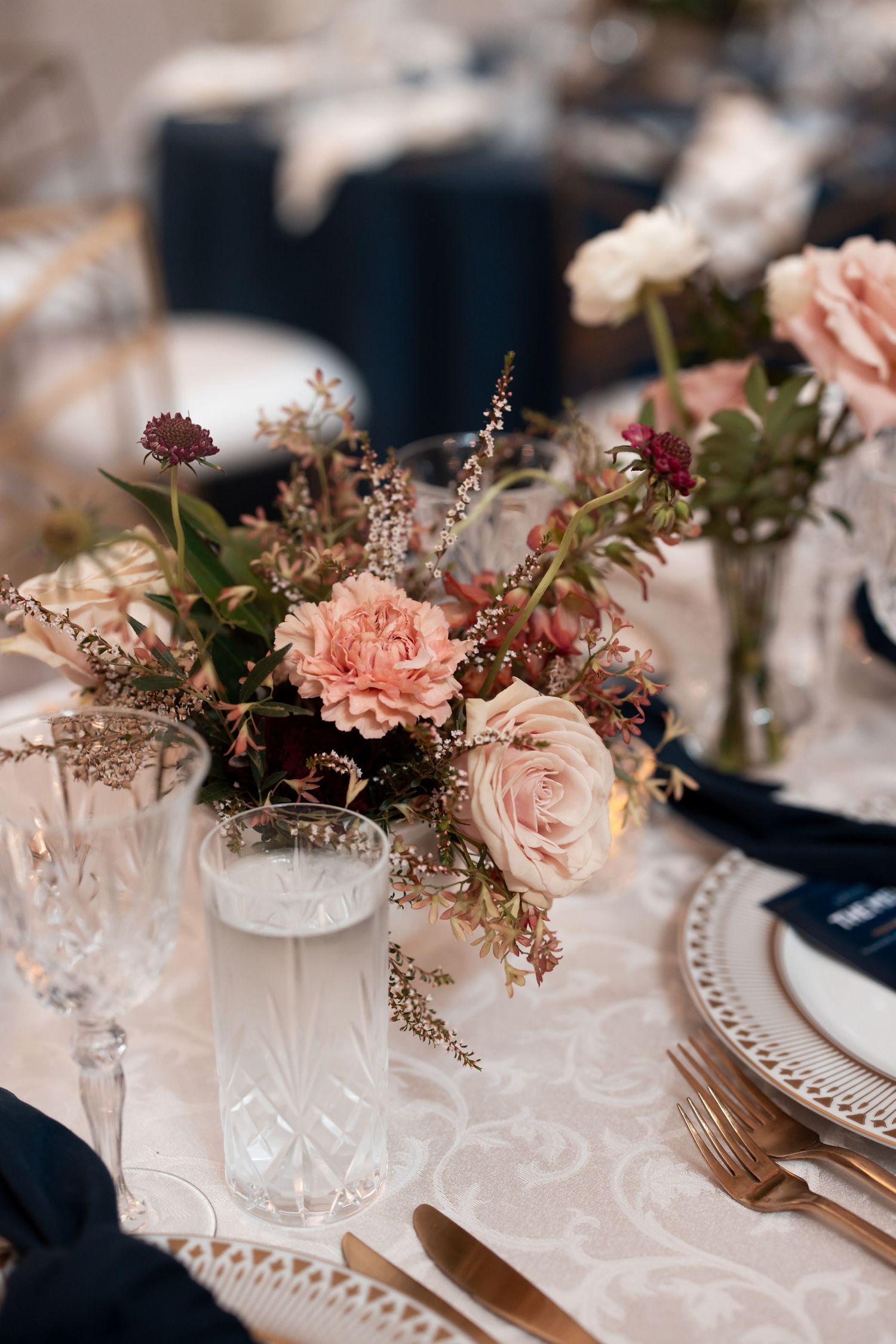 Elegant wedding table setting with blush pink roses, carnations, crystal glassware, navy napkins, and gold cutlery on white lace.