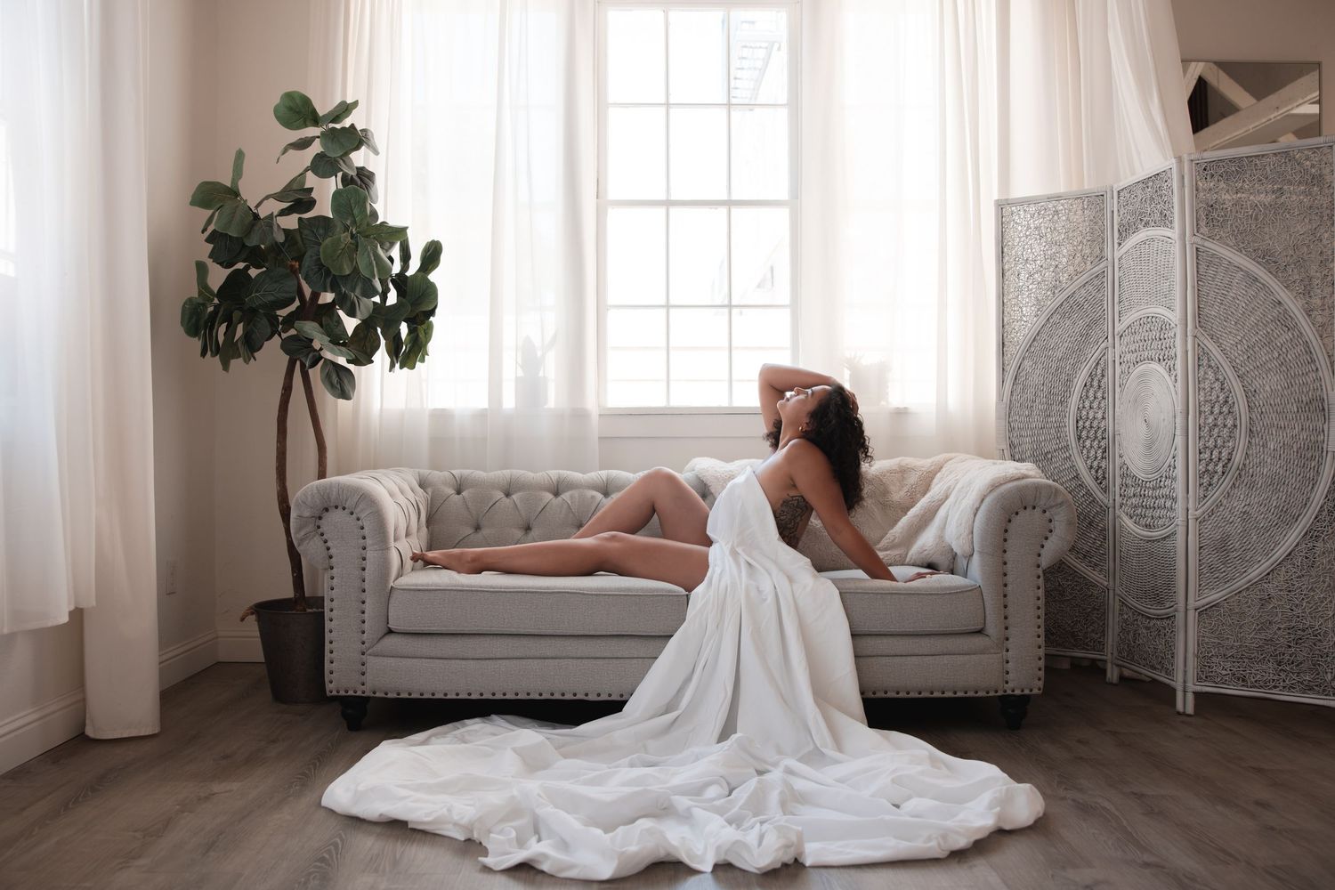 A figure in a flowing white gown reclines elegantly on a gray tufted sofa near a potted fiddle leaf fig and decorative screen.