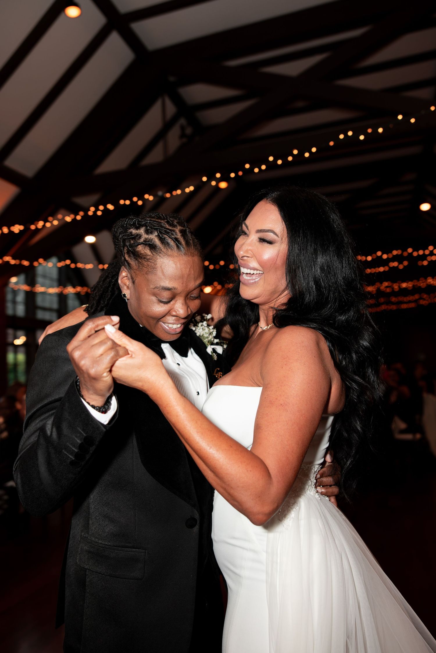 Two brides laugh and dance together at their wedding reception, one in a black tuxedo and one in a white gown, under string lights.