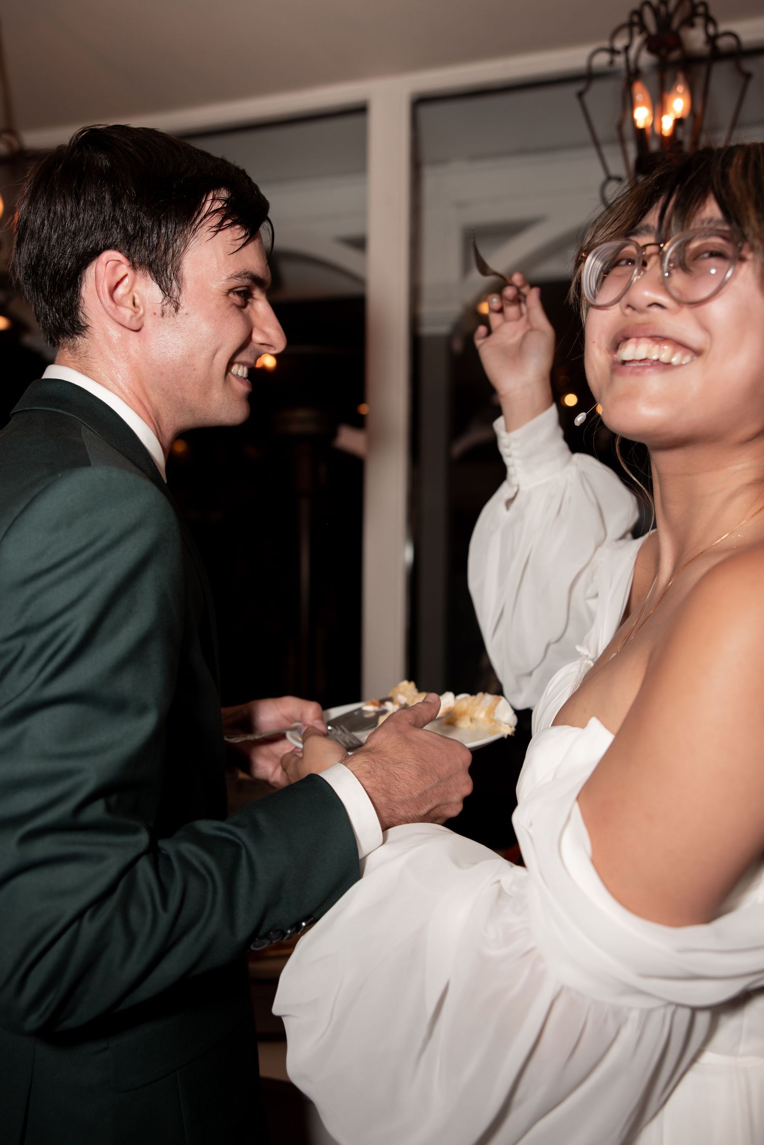 A couple laughs and dances together at a wedding reception, with one person wearing a green suit and another in white.