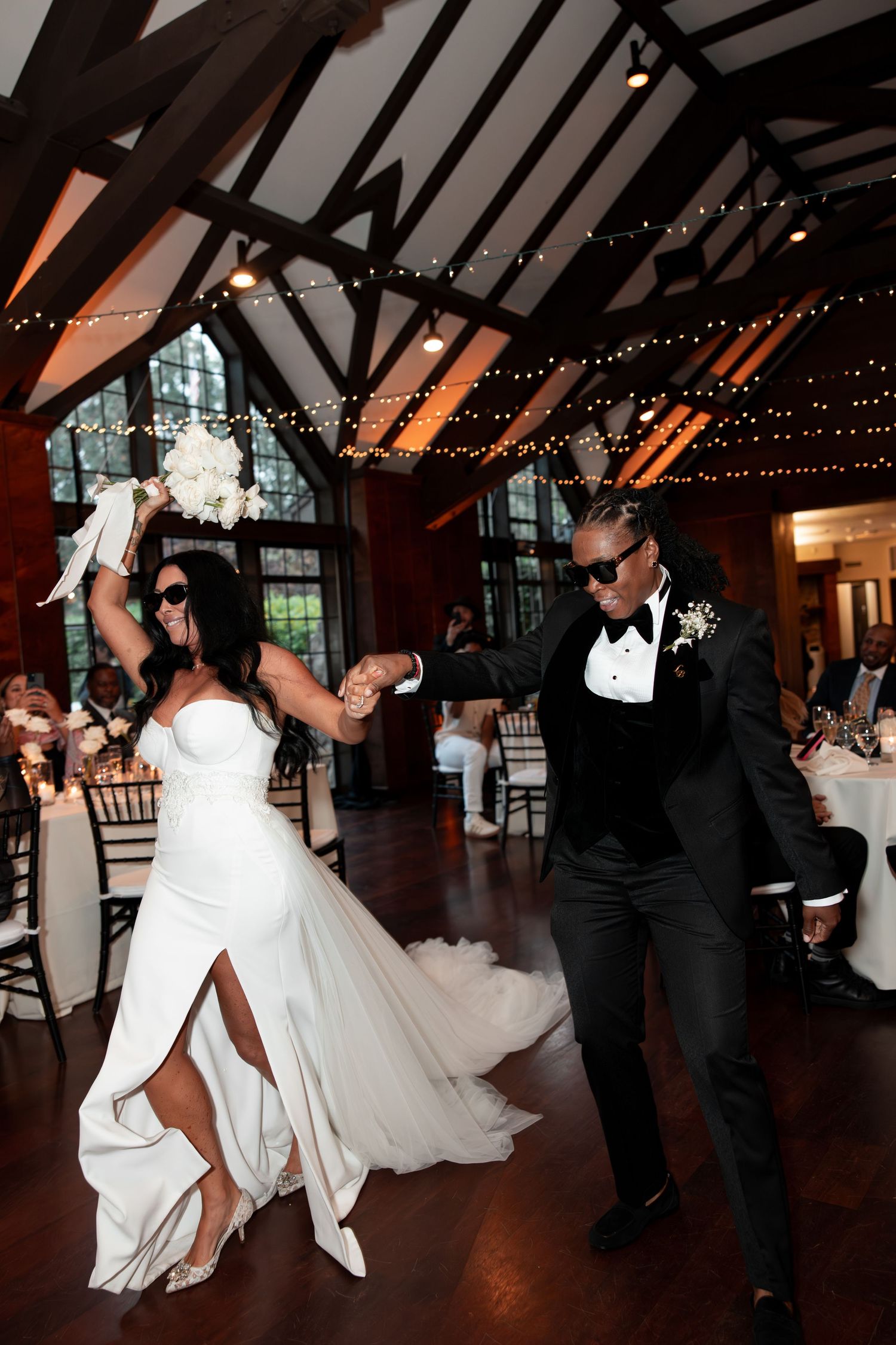 A couple dances together at their wedding reception in a rustic venue with exposed beams and twinkle lights.