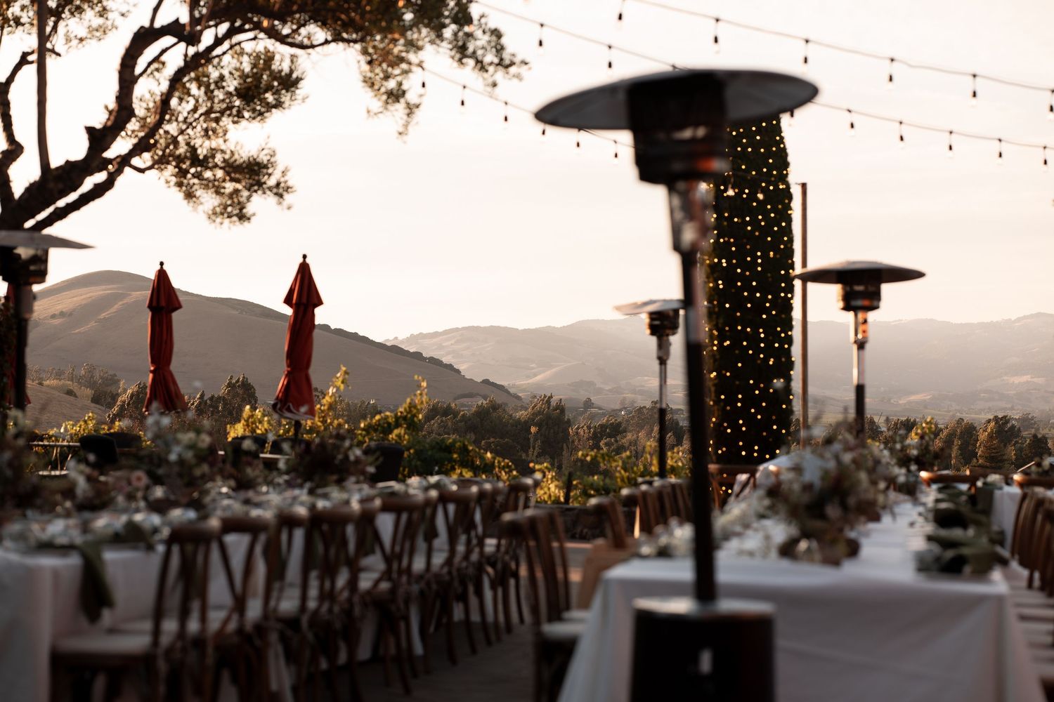 A scenic outdoor wedding reception setup at sunset with string lights and mountain views in the background.
