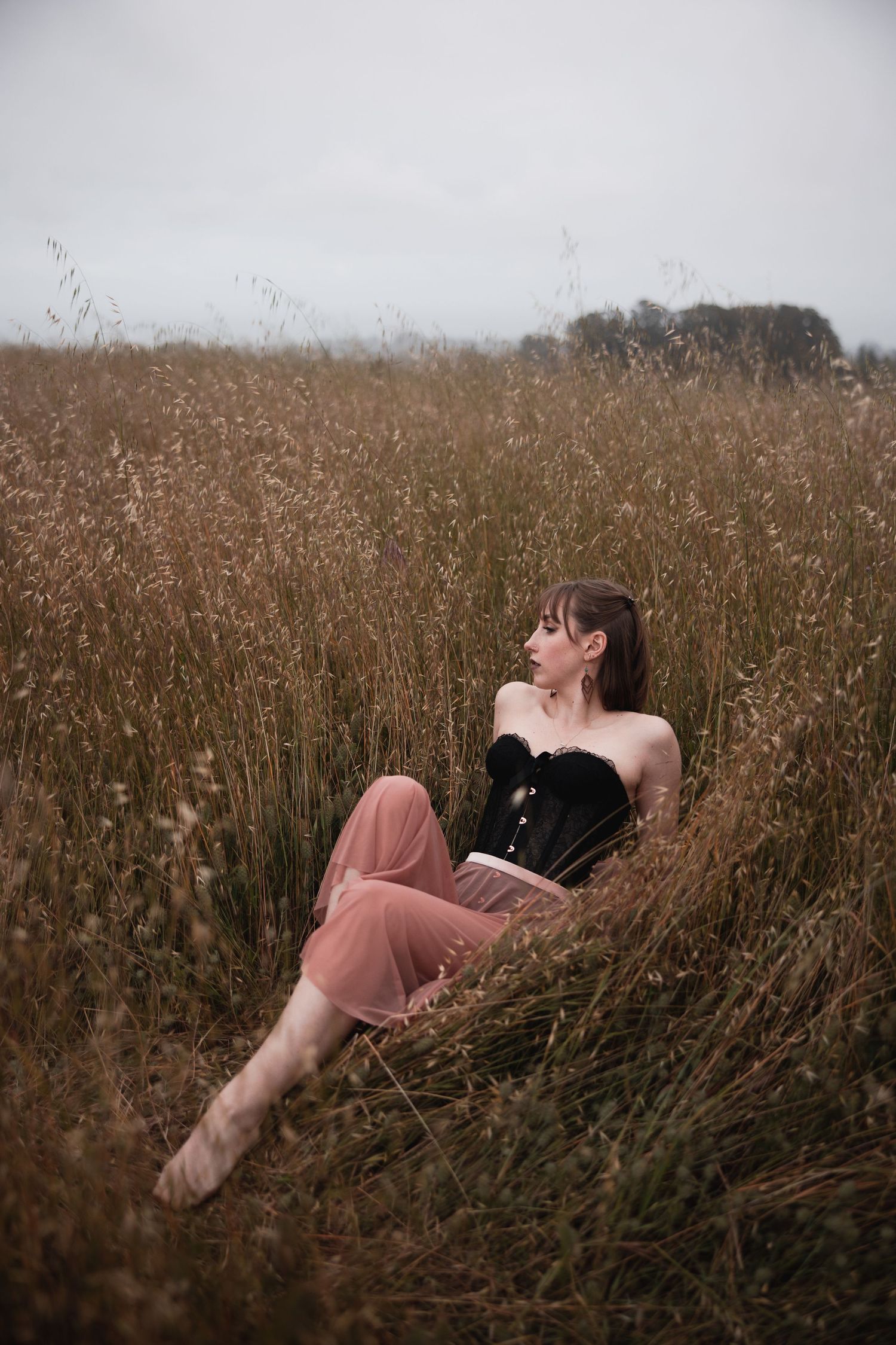 A dreamy portrait in a golden wheat field shows a figure in a black corset and pink skirt reclining among tall grass.