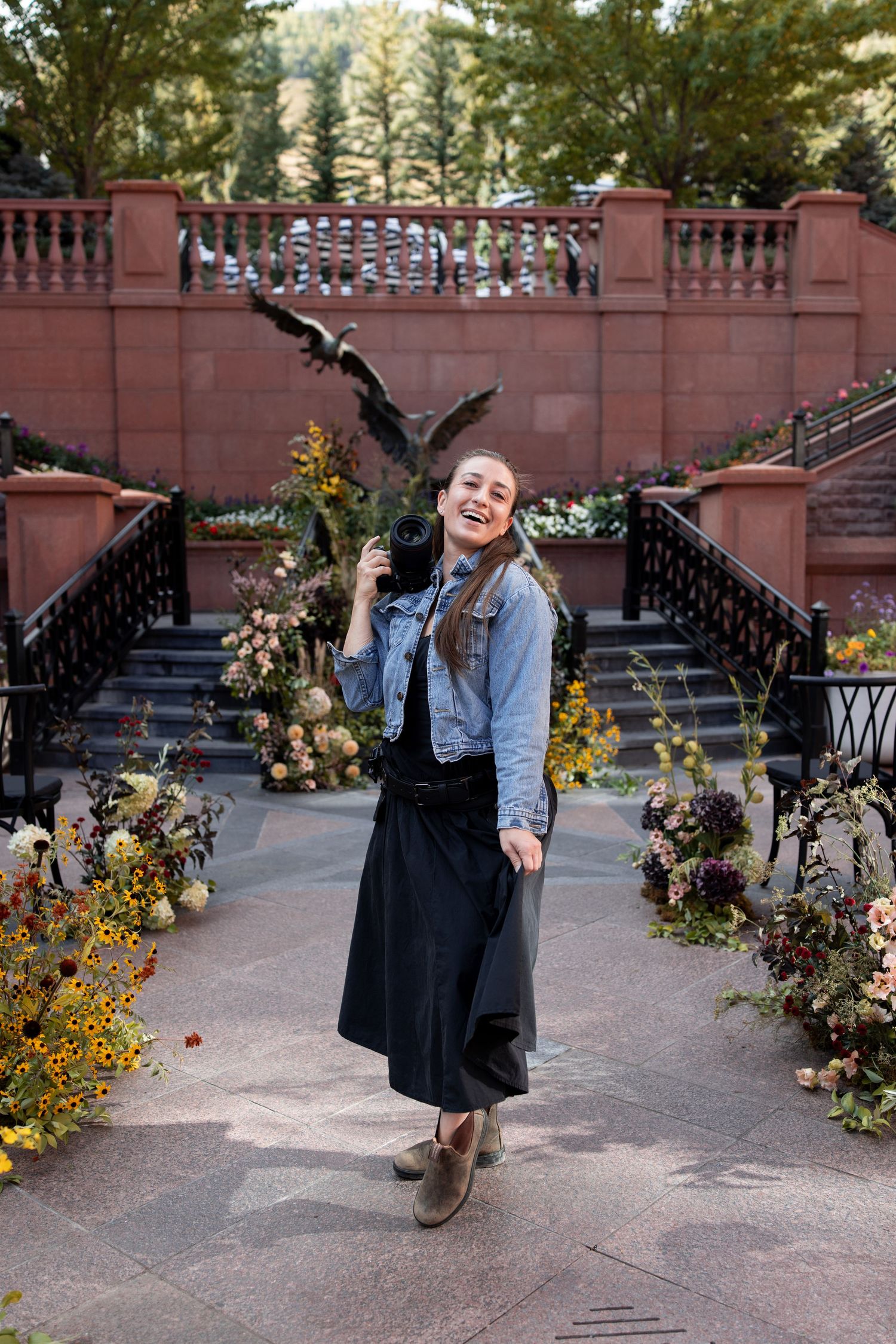 Smiling woman in denim jacket and black skirt poses outdoors near floral arrangements and a bronze eagle sculpture.