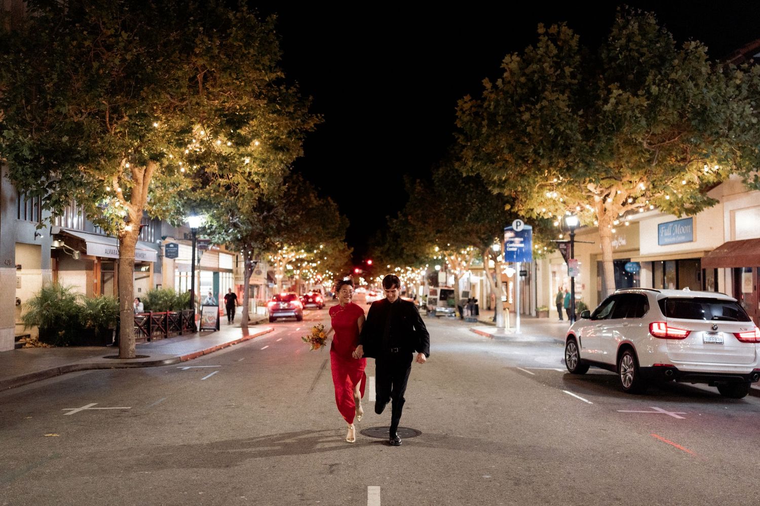 A nighttime street scene with illuminated trees and parked cars along a commercial district sidewalk.
