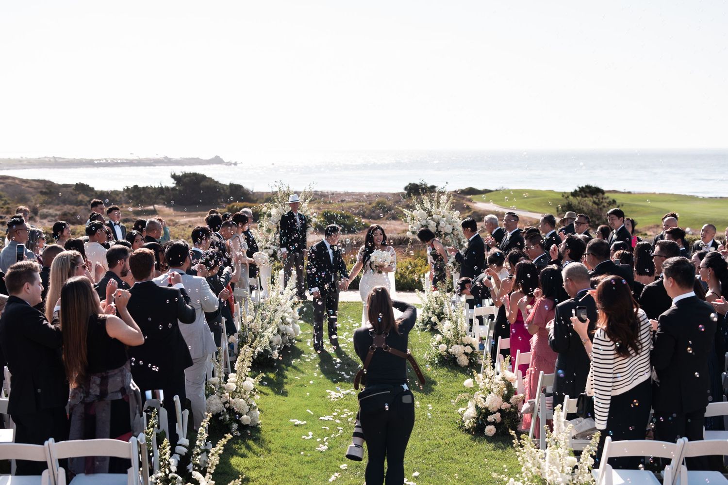 A large crowd gathers outdoors at a coastal ceremony with people standing in rows facing the ocean.