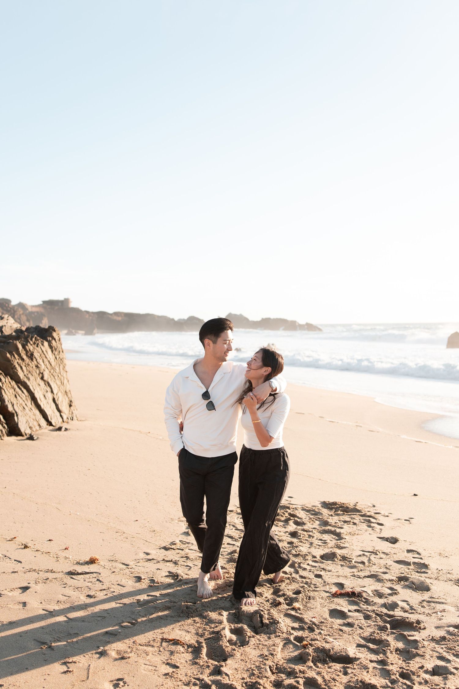 A couple walks hand in hand on a sunny beach, smiling at each other in matching white tops and black pants.