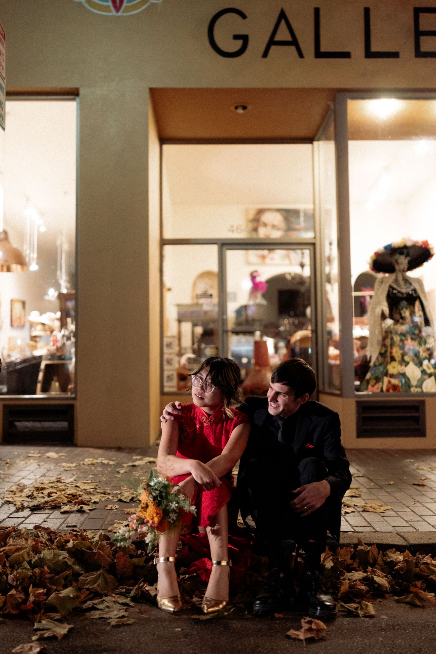 A couple sits on a bench in front of an illuminated gallery storefront with autumn leaves scattered on the ground.