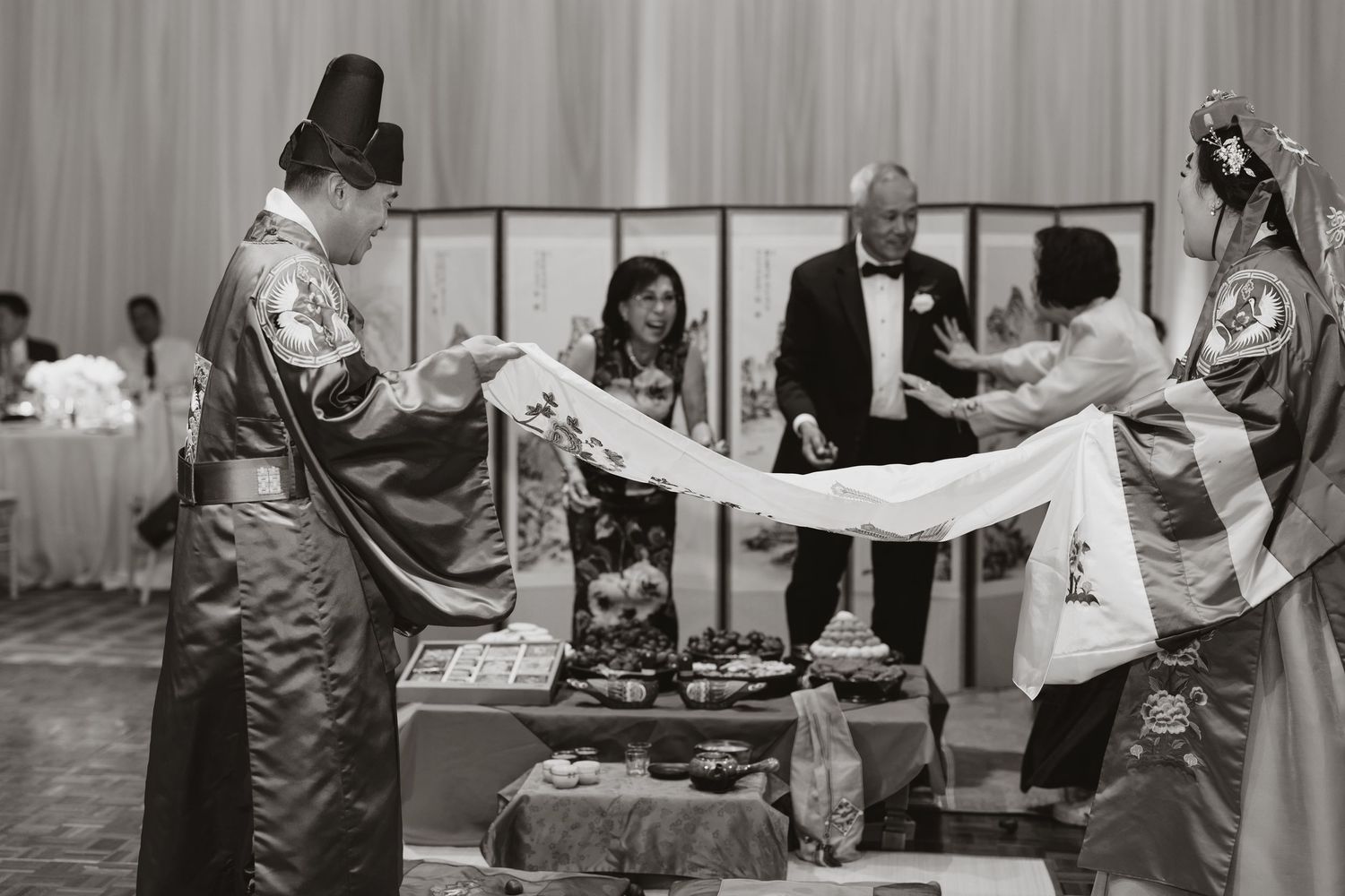 Black and white photo of a traditional Korean wedding ceremony with guests in hanbok attire holding a white cloth.