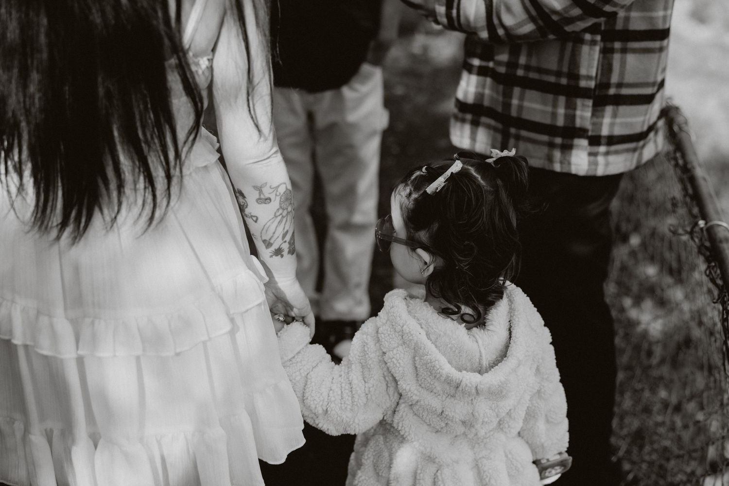 A black and white photograph shows a small child holding hands with adults while wearing a white ruffled dress.