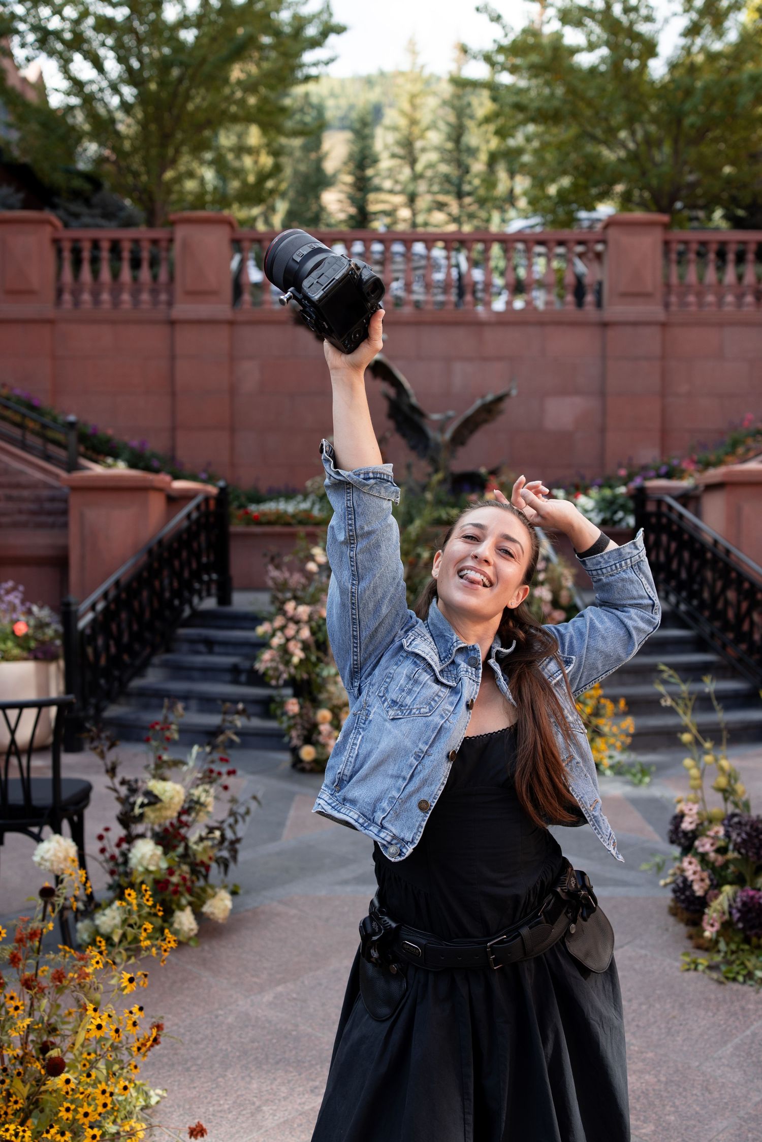 A joyful female photographer raises her camera triumphantly outdoors near floral arrangements and stone stairs.