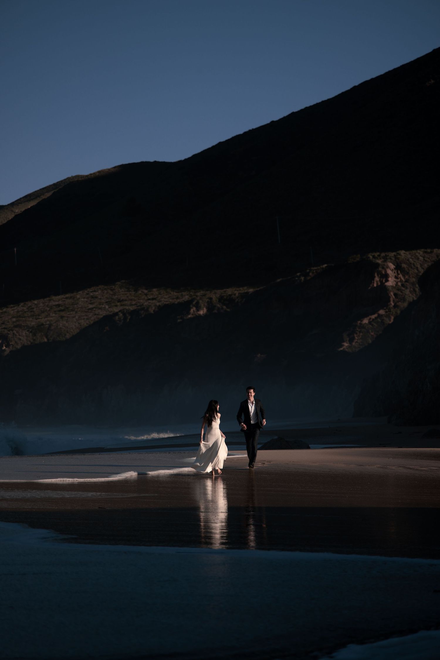 A couple walks along a dramatic black sand beach at sunset with mountains in the background.