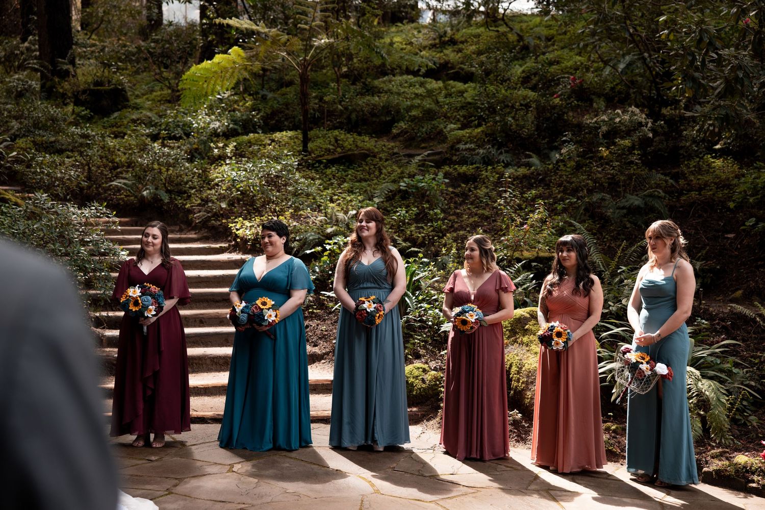 A wedding party in burgundy and teal dresses poses outdoors on stone steps surrounded by lush greenery.