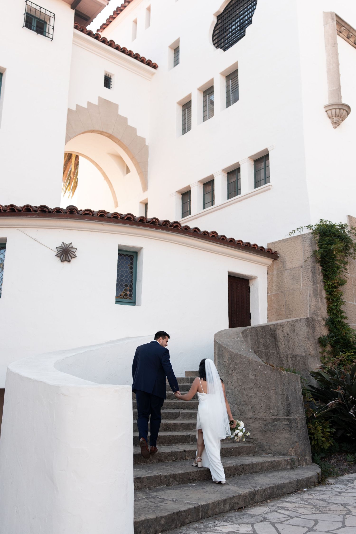 Two people walk up stone steps at a Mediterranean-style white stucco building with arched doorways and terracotta roof tiles.